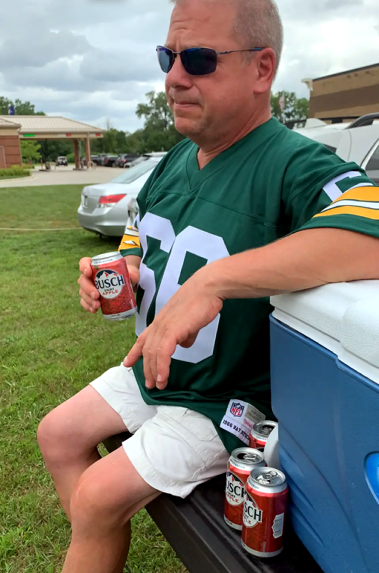 Man in green football jersey (number 86) wearing sunglasses sits by a blue cooler, holding a Busch Apple can in a parking lot with other cans nearby.