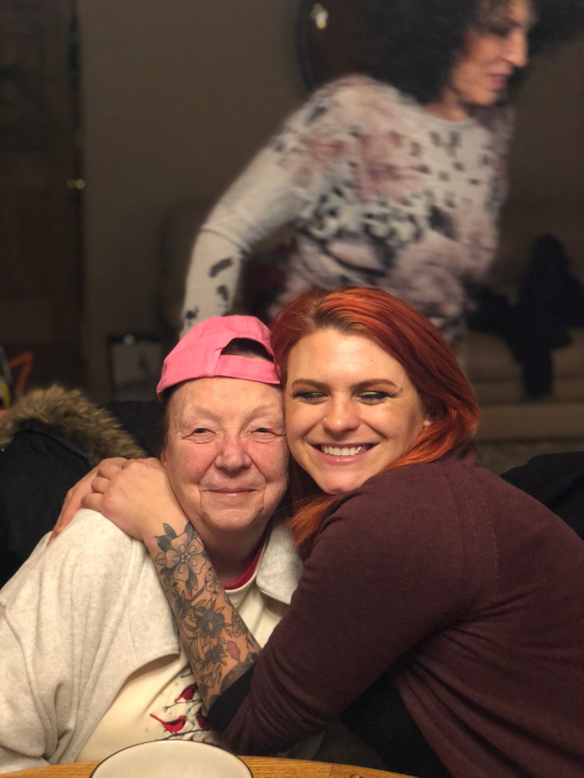 Smiling woman with red hair in a hug with an older man wearing a pink cap at a table, tattooed arm visible.