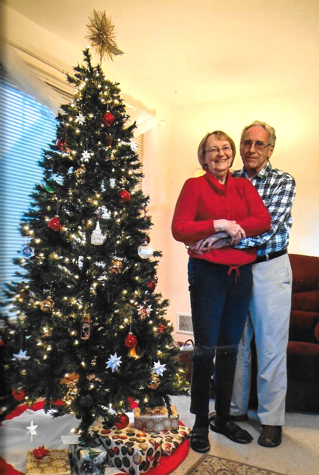 Older couple embracing beside a decorated Christmas tree with lights and gifts underneath.