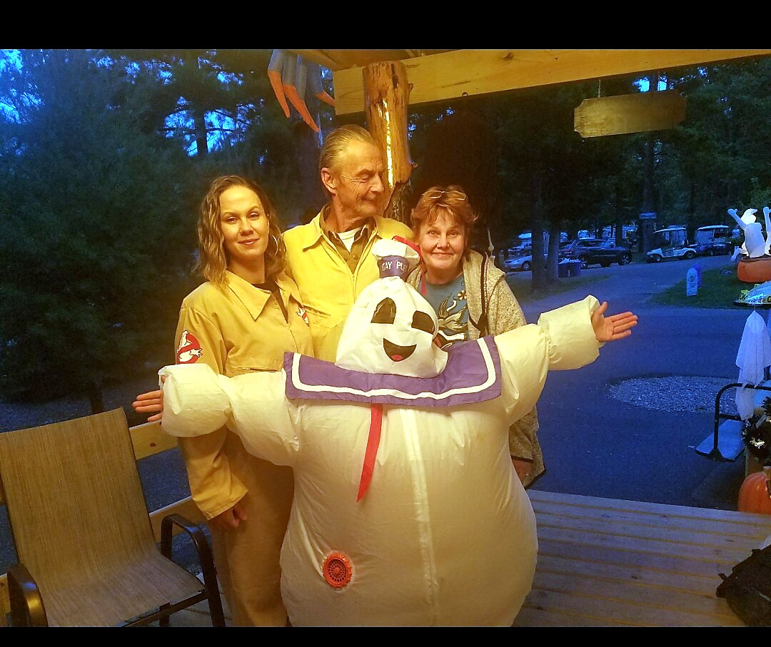 Two people in Ghostbusters-style uniforms pose with a person wearing a large inflatable Stay Puft Marshmallow Man costume on a wooden porch at dusk, outdoors.