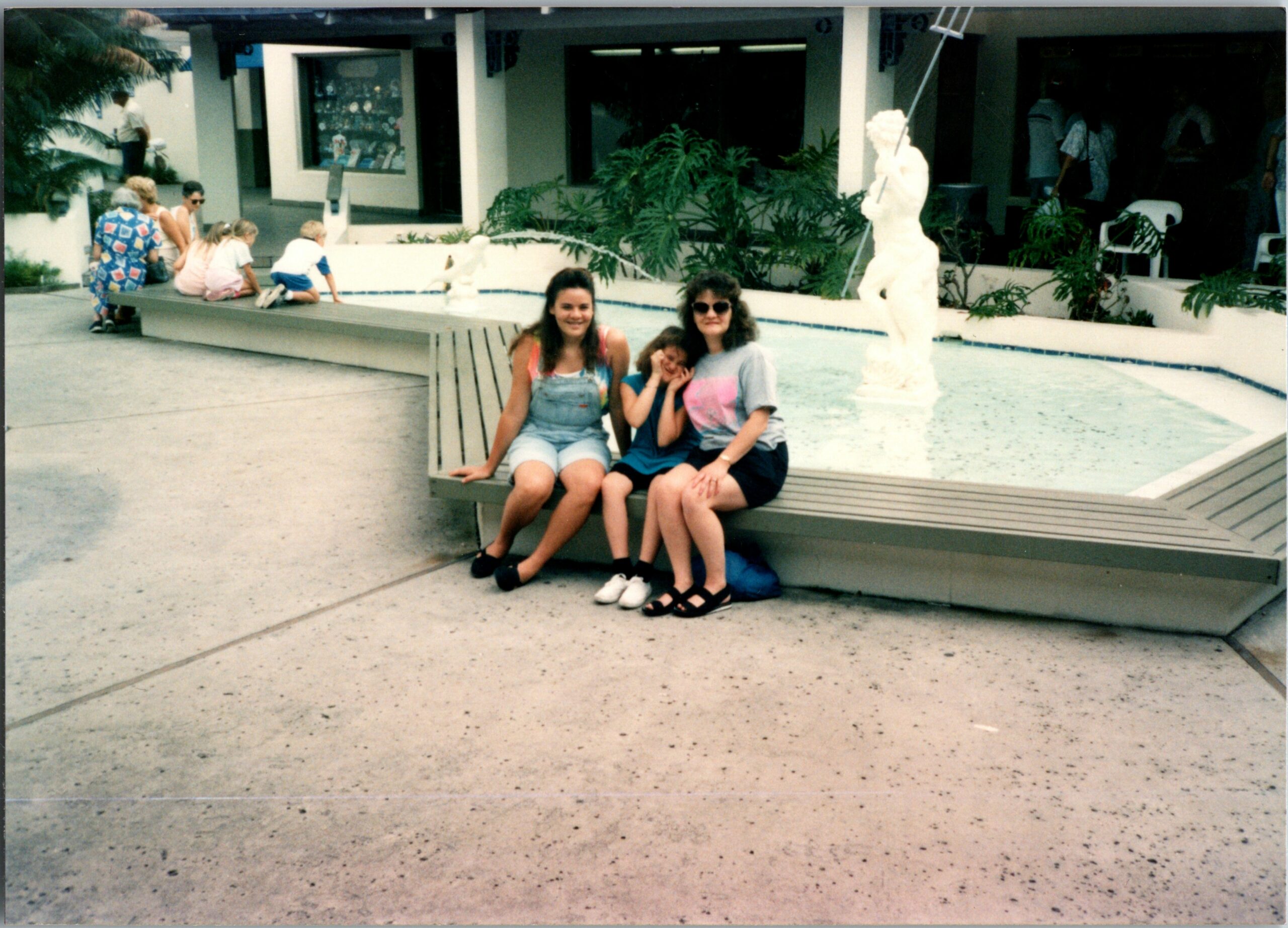 Three girls sit on a wooden bench beside a shallow fountain, smiling at the camera with a statue and greenery behind them.