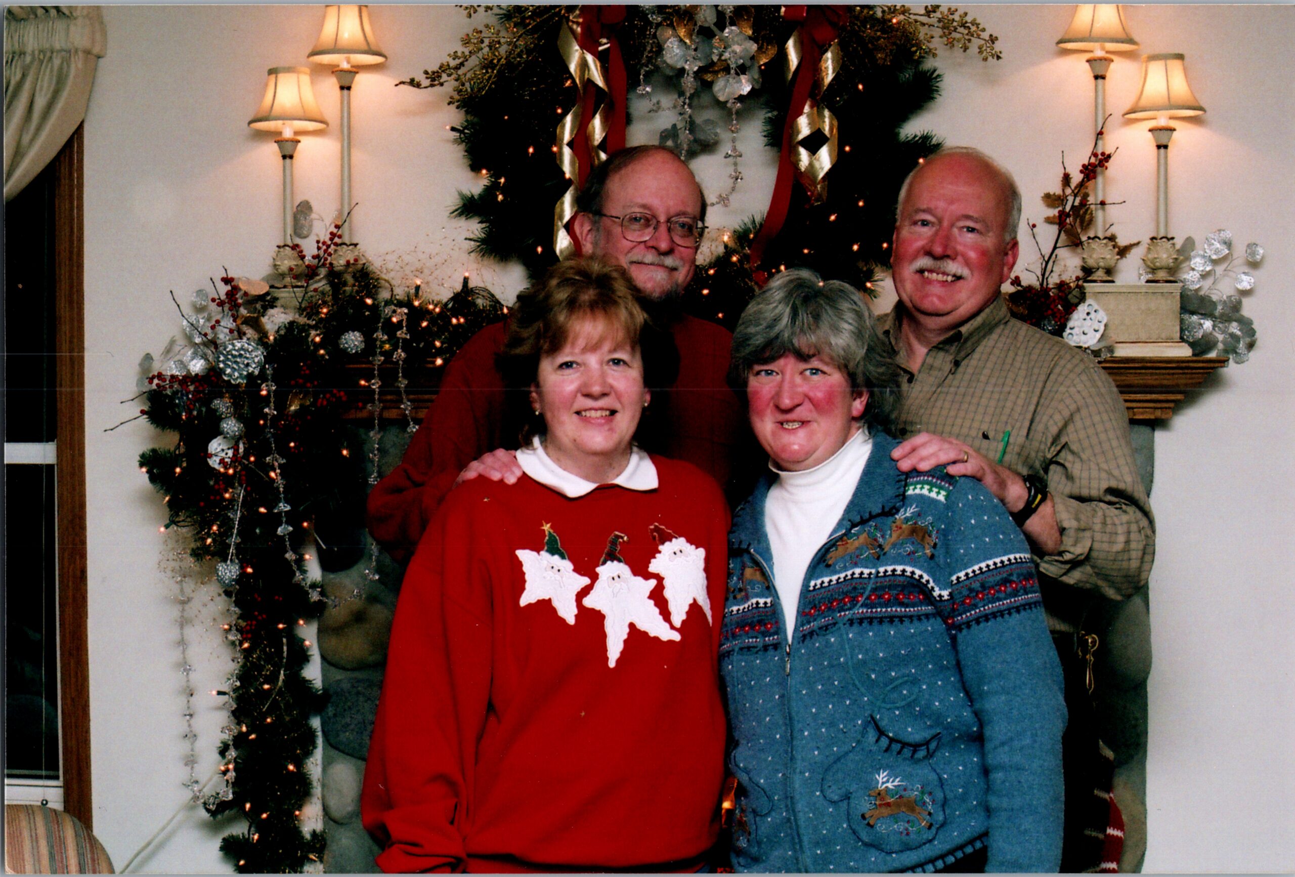 Four adults in festive sweaters pose for a Christmas photo in front of a decorated tree and garlanded mantelpiece.