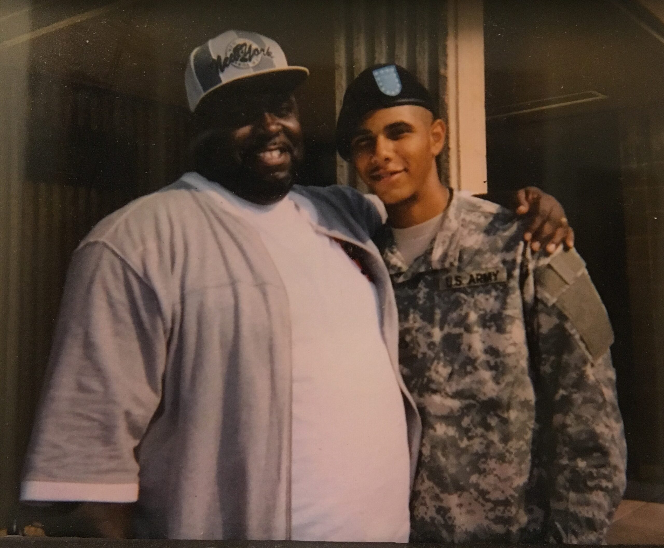 Two men pose for a casual photo: a smiling man in a gray shirt and baseball cap with another man in Army camouflage, arm around each other.