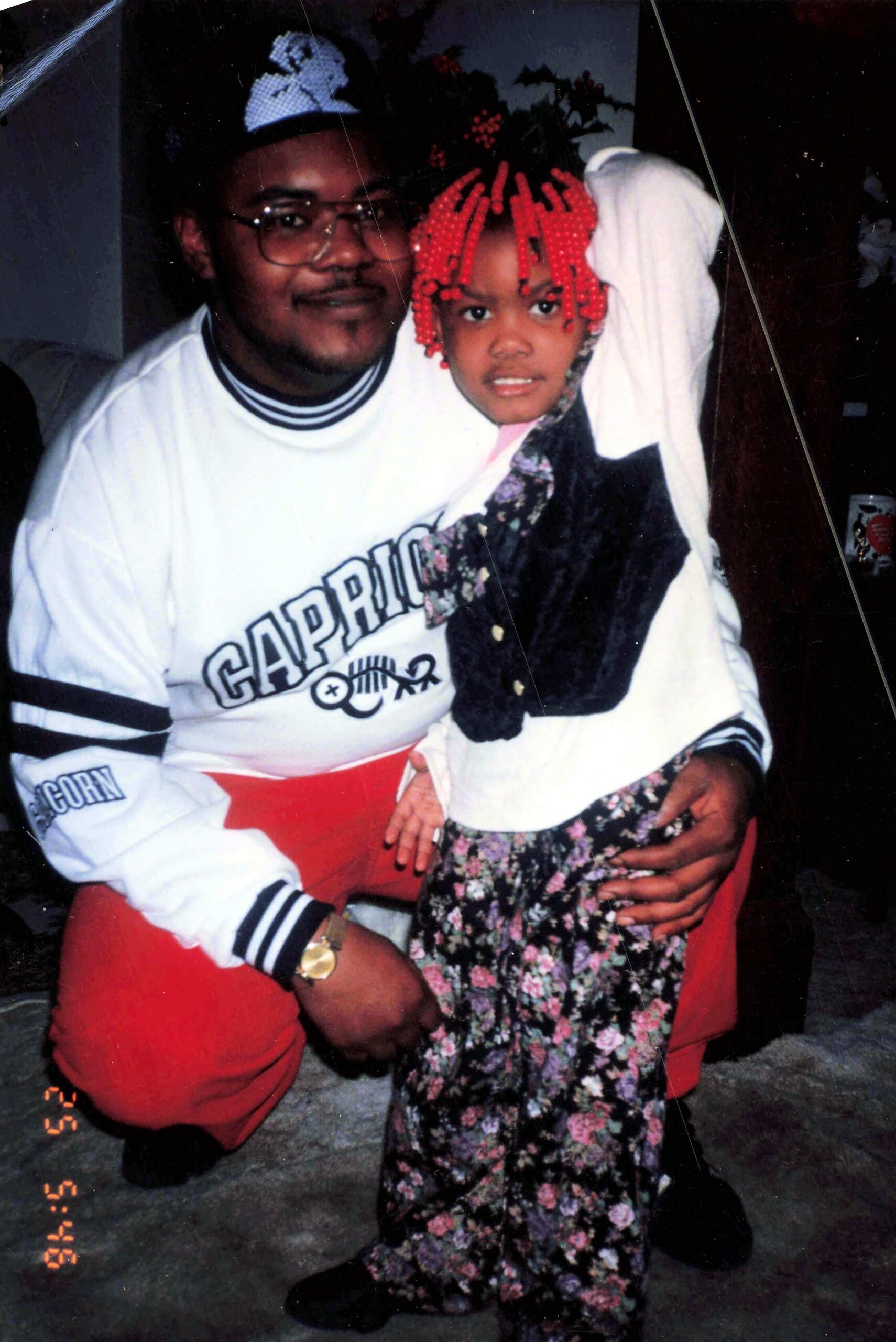 Man wearing glasses and a white striped-sleeve sweatshirt sits beside a girl with red beaded hair; they pose closely indoors with a floral dress on the girl and red pants on the man beside her.