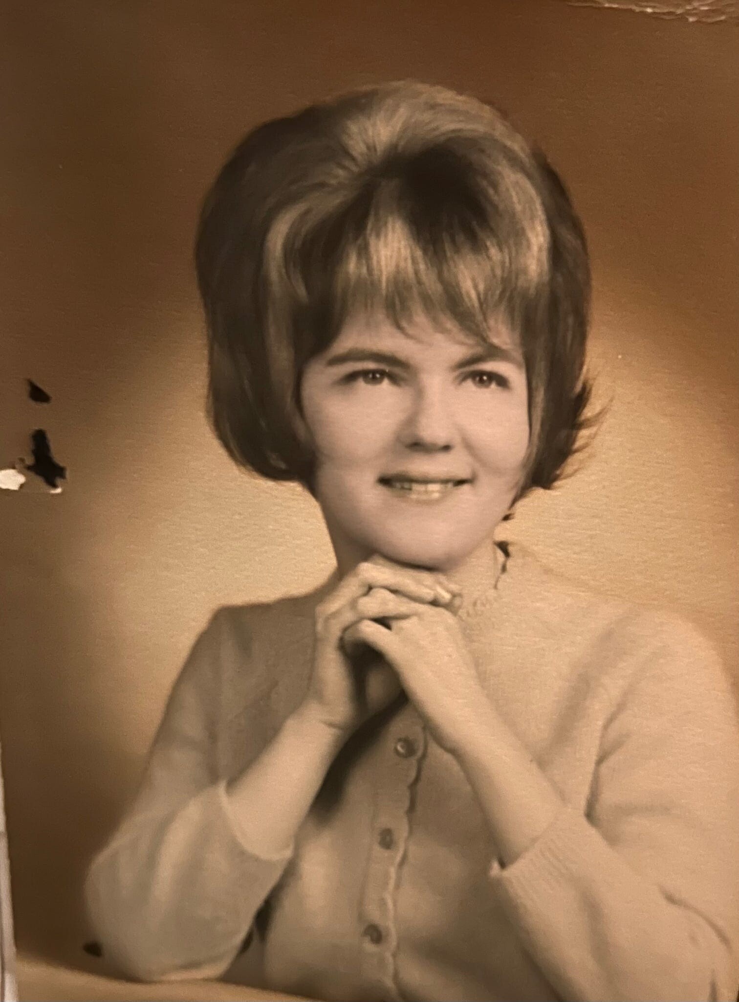 Black-and-white portrait of a young woman with a voluminous 1960s hairstyle, smiling with hands clasped under her chin.