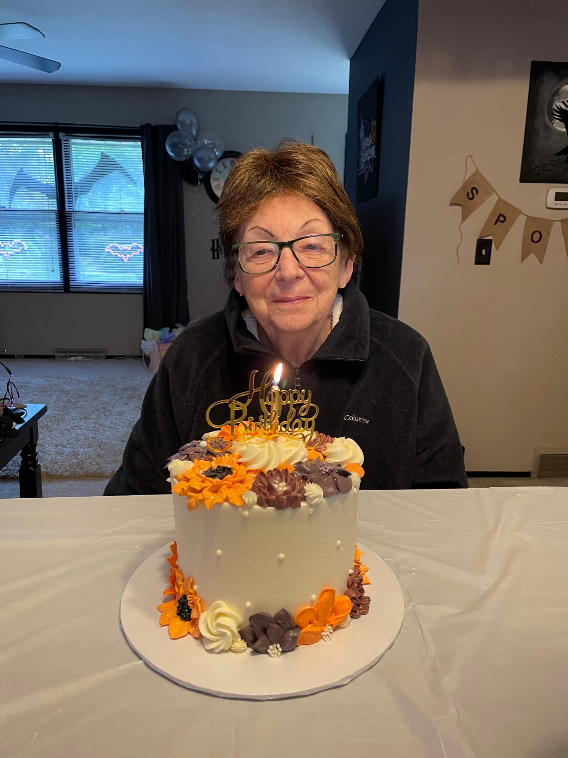 Older woman in glasses birthdaying a tiered cake with orange, white, and lavender frosting decorations tabbed around the edges.