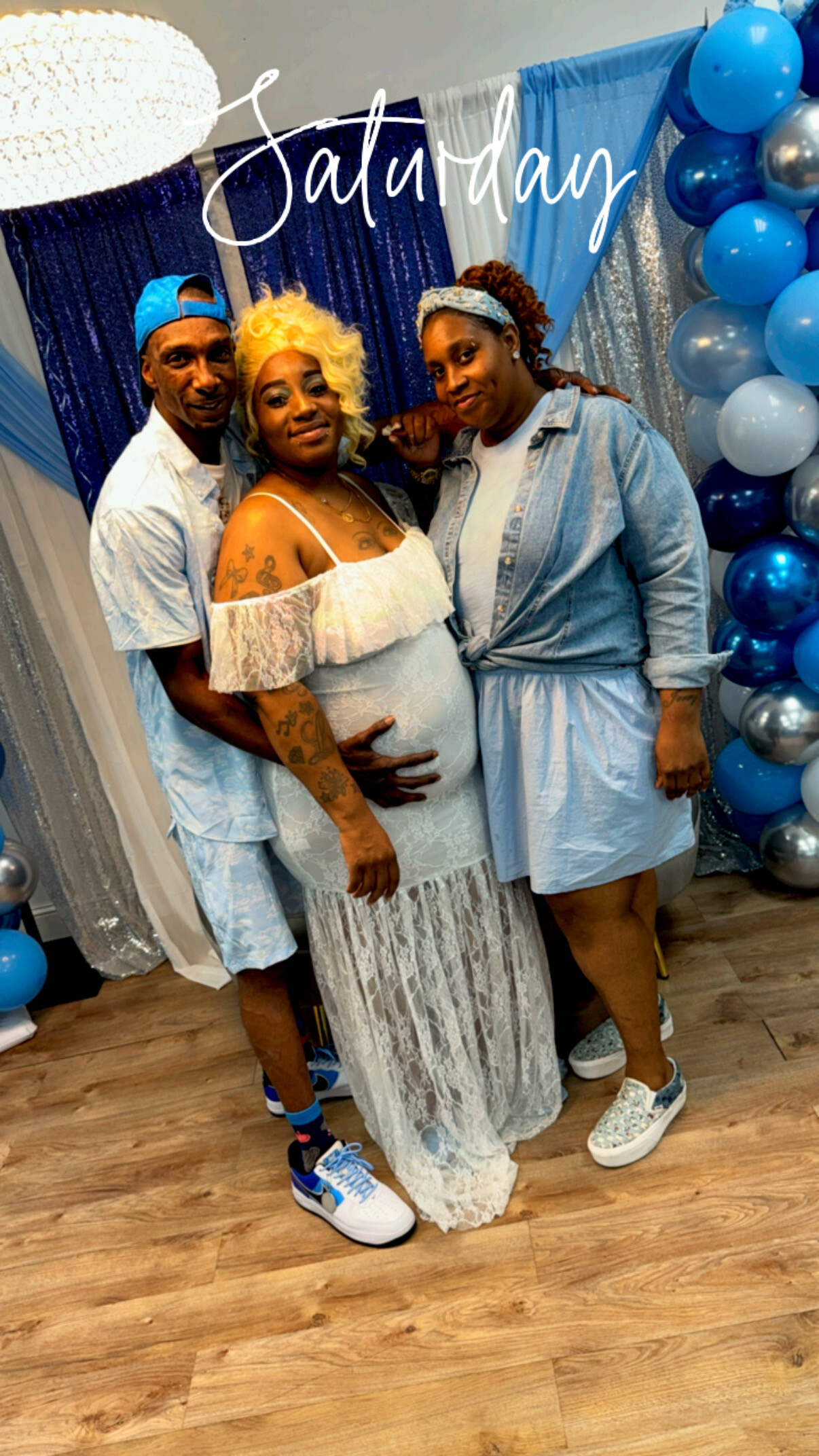 A pregnant woman in a white lace dress stands with a man and a woman at a blue-and-silver balloon backdrop for a Saturday event.