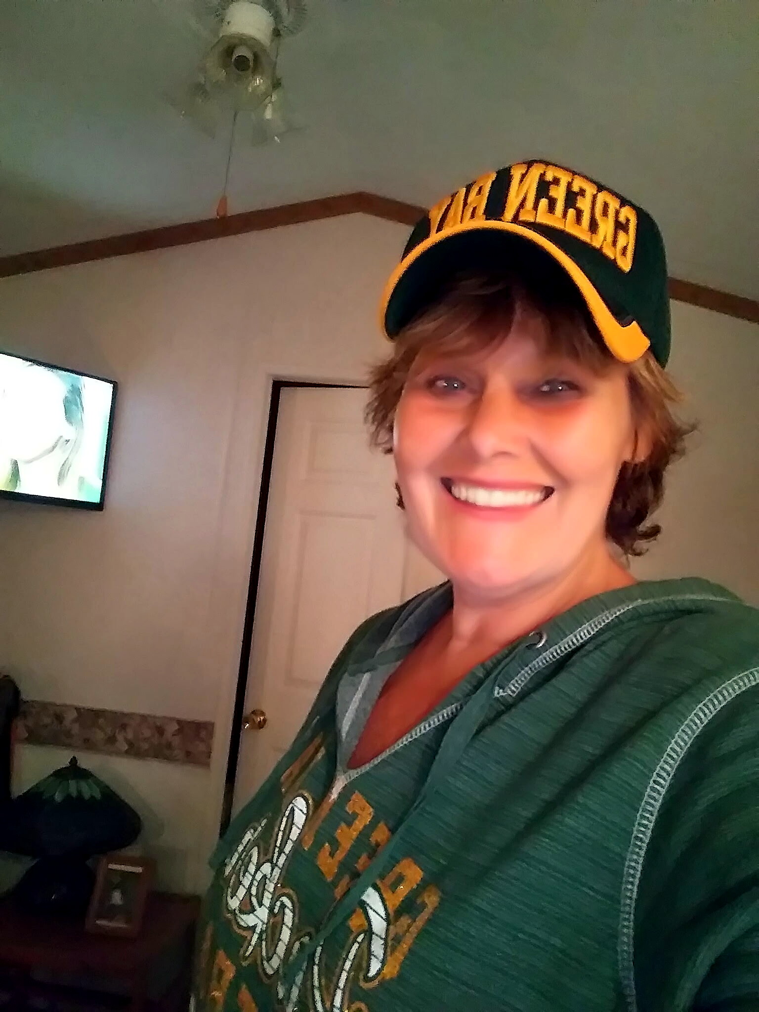 Smiling person wearing a green hoodie and a green-yellow cap, taking a selfie indoors with a TV in the background.