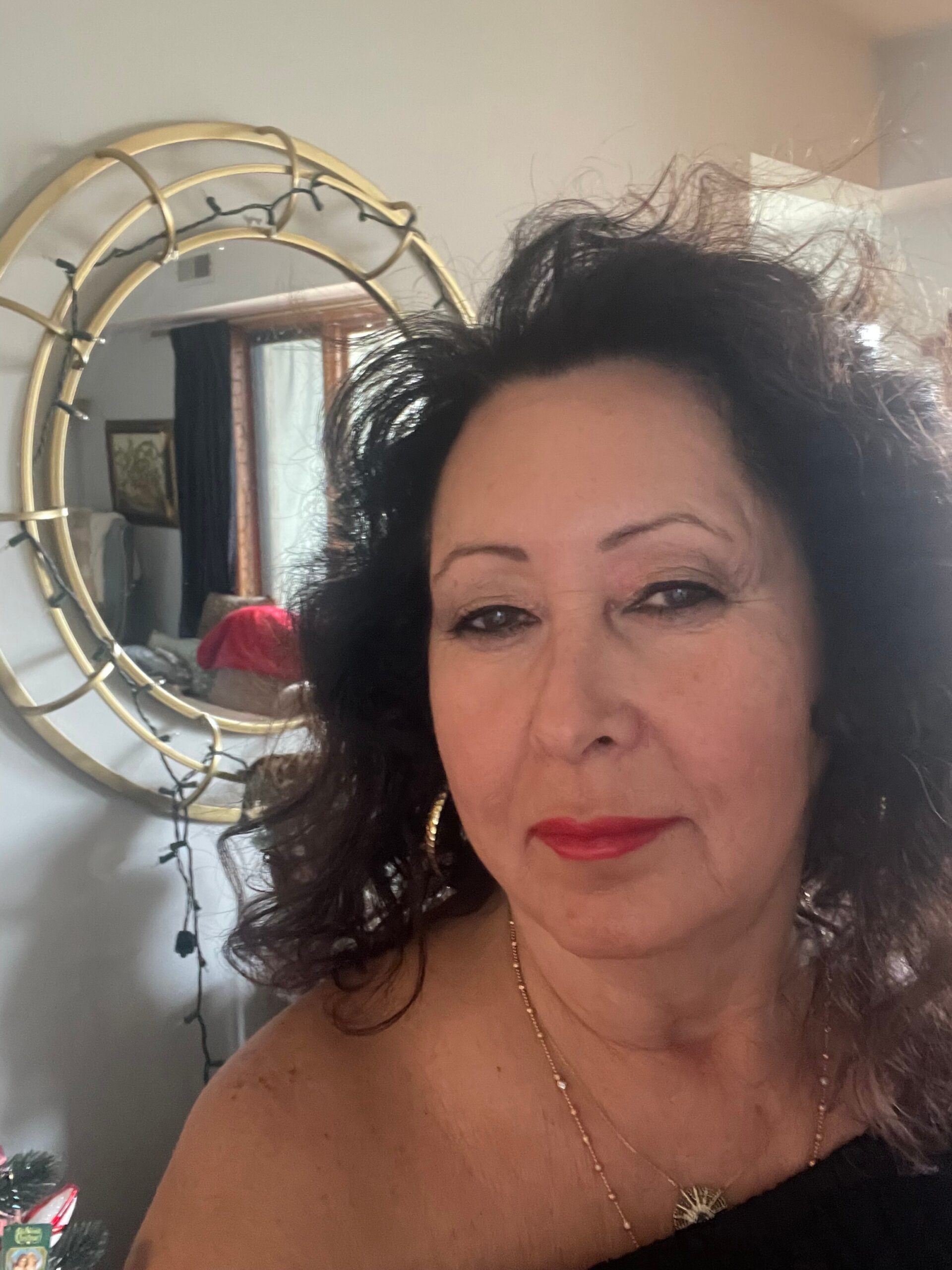 Close-up portrait of a woman with dark hair, red lipstick, and a gold necklace, reflected in a round wicker-framed mirror behind her.