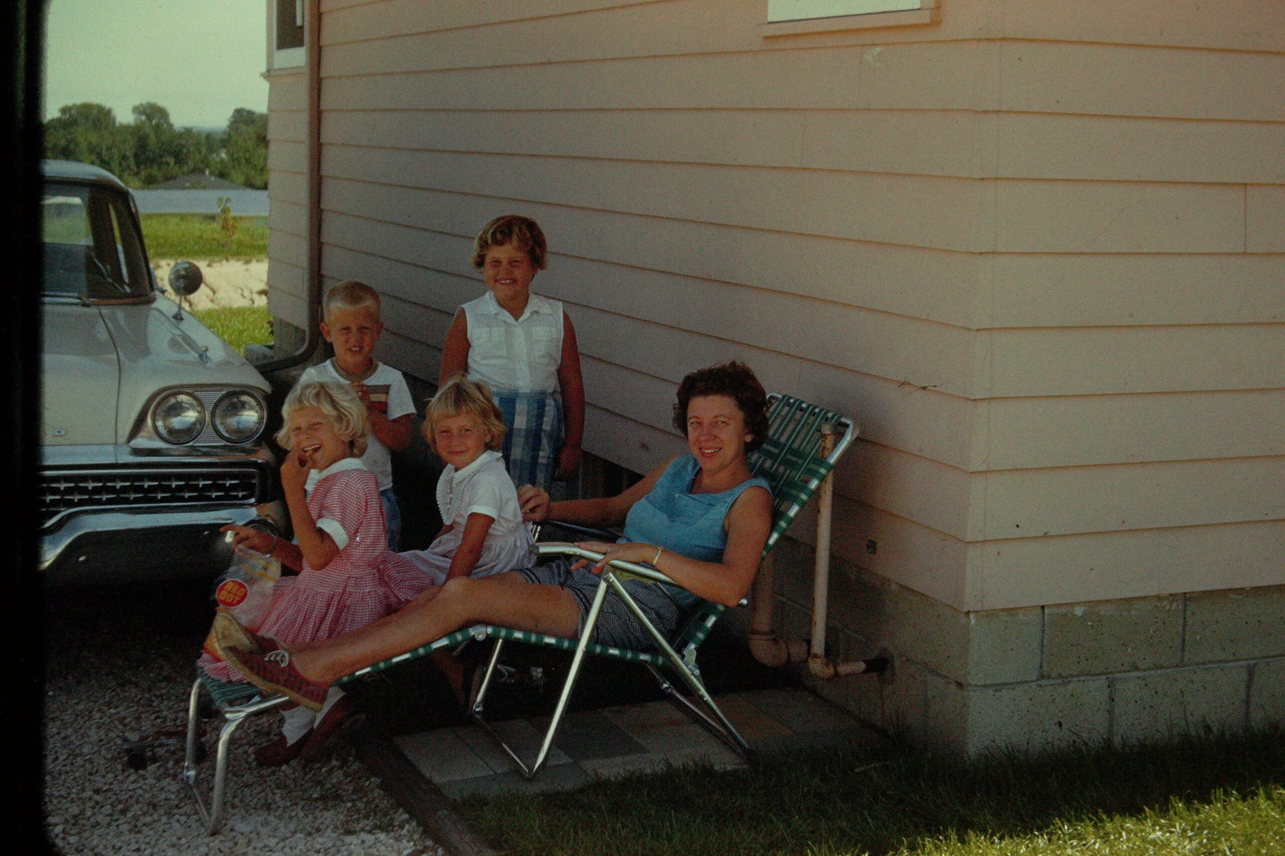 A family gathering outside a beige siding house, seated in lawn chairs beside a vintage white car.