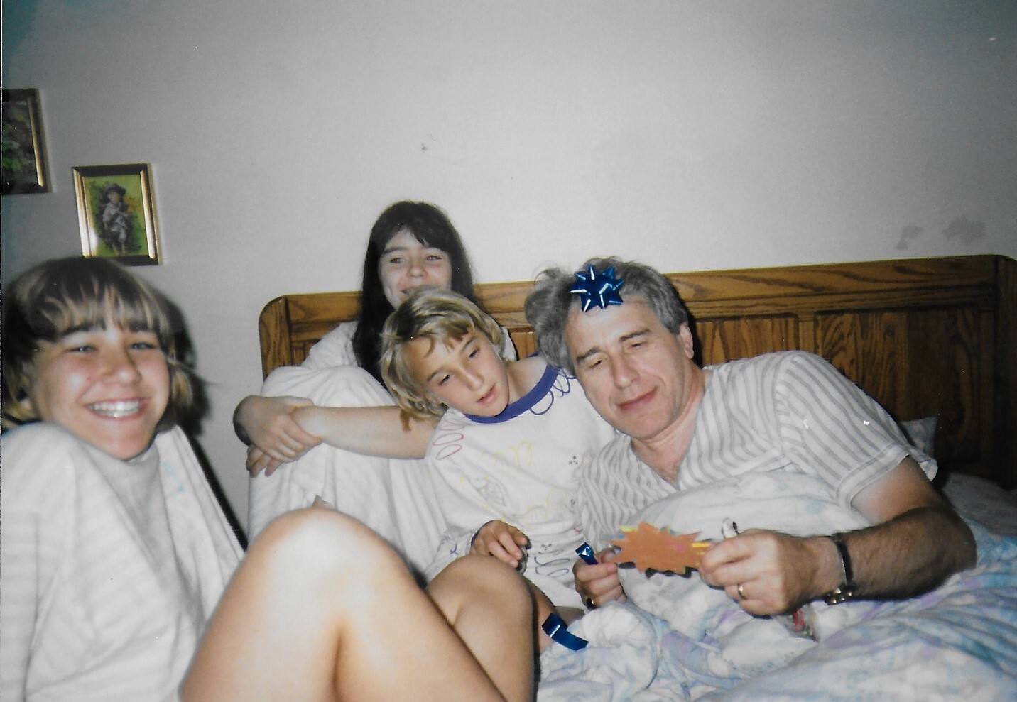Four people sit and lean on a bed in pajamas, smiling for a casual photo in a cozy bedroom.