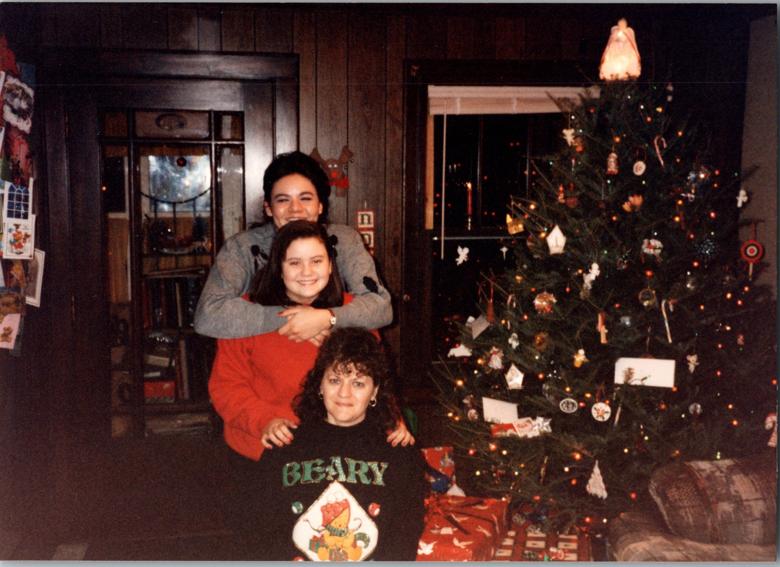 Three people posing for a Christmas photo: two adults behind a child, standing beside a decorated Christmas tree with gifts underneath in a cozy home setting.