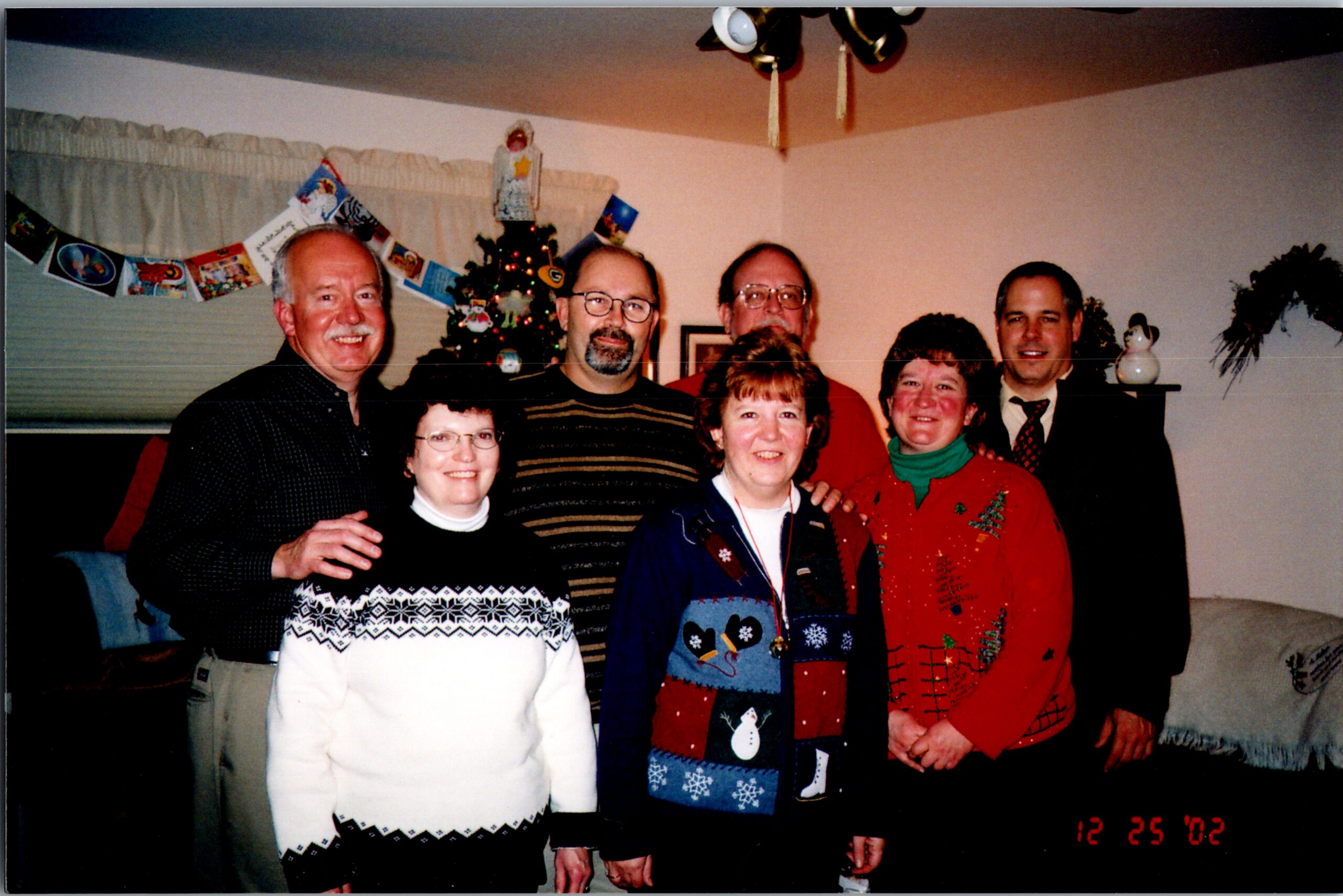 Six adults in festive sweaters pose for a Christmas photo indoors, with a decorated tree in the background.