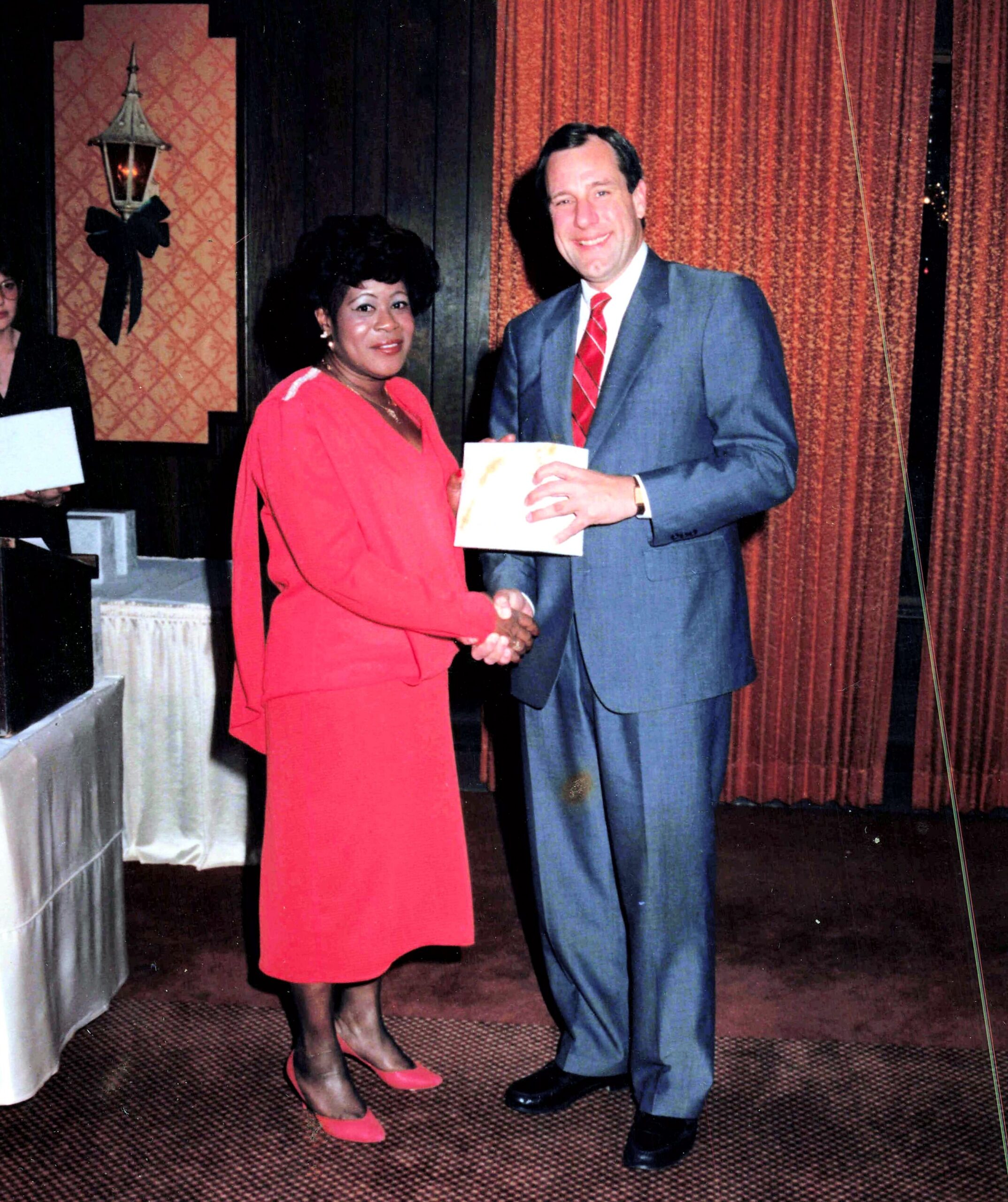 Man in a gray suit with a red tie shakes hands with a woman in a red dress on a stage, handing her a certificate envelope.