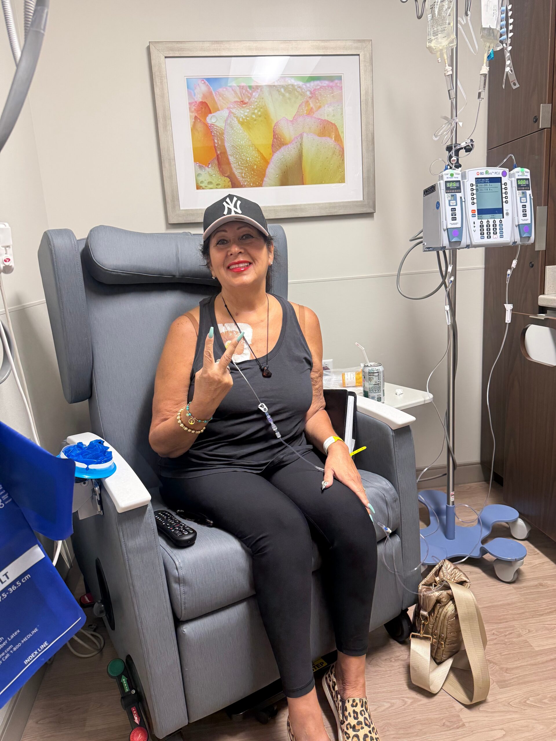 Smiling woman in a clinical chair giving a peace sign with an IV line attached nearby.