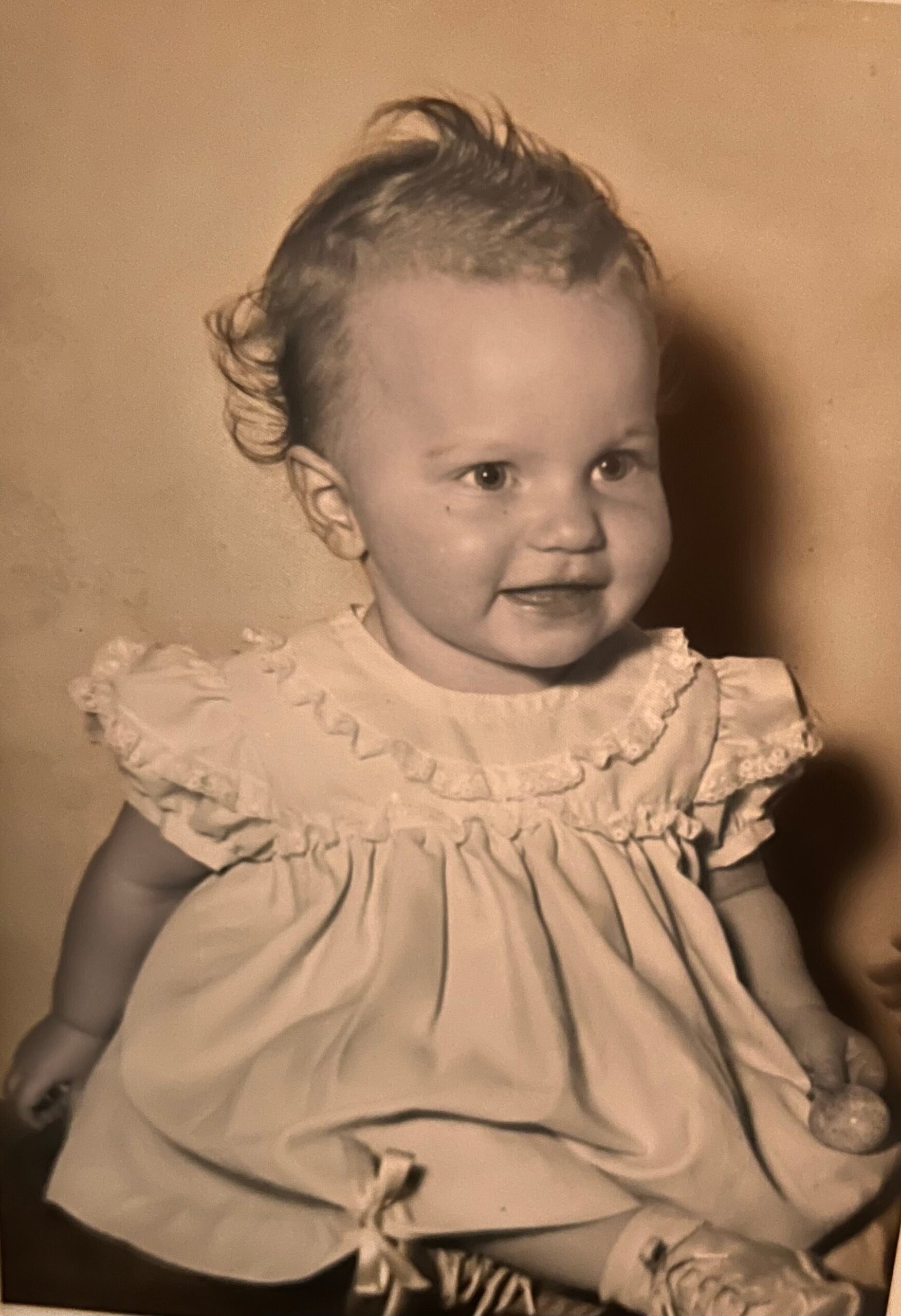 Black-and-white photo of a smiling baby wearing a frilly, lace-trimmed dress and short curls, facing slightly to the right.