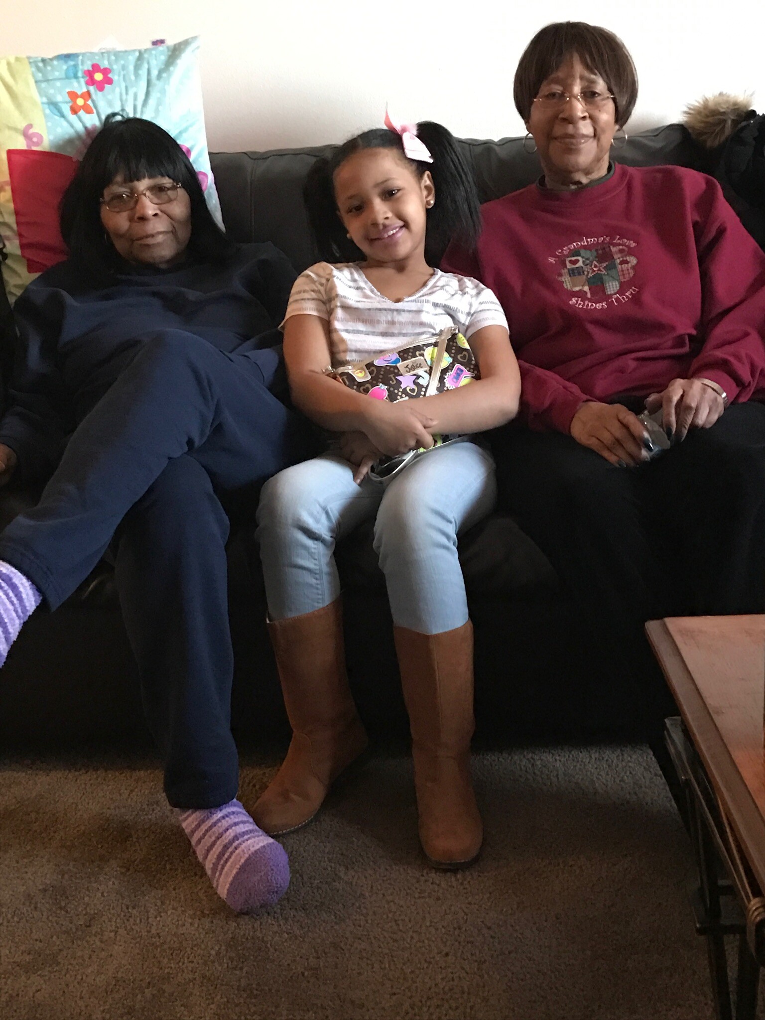 Three generations sit on a couch: grandmother on left in dark clothing, a smiling young girl with a pink bow at center holding a colorful gift bag, grandmother on the right in a maroon sweatshirt.