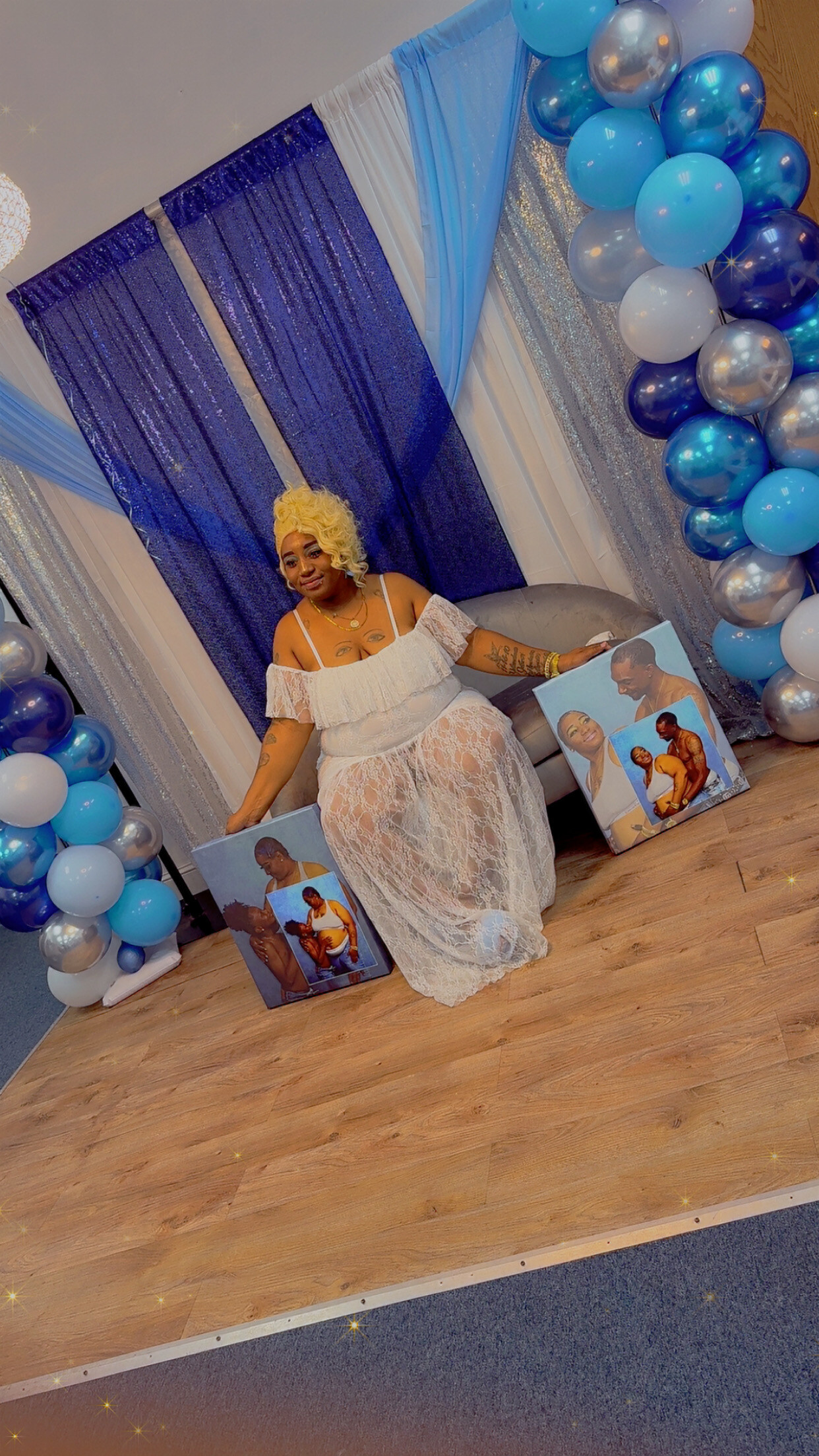 Woman with blonde curly wig in a white lace off-shoulder dress sits on a sofa beneath a blue-silver event backdrop, surrounded by a blue, white, and silver balloon arch; several canvases with family photos lean on the floor nearby.