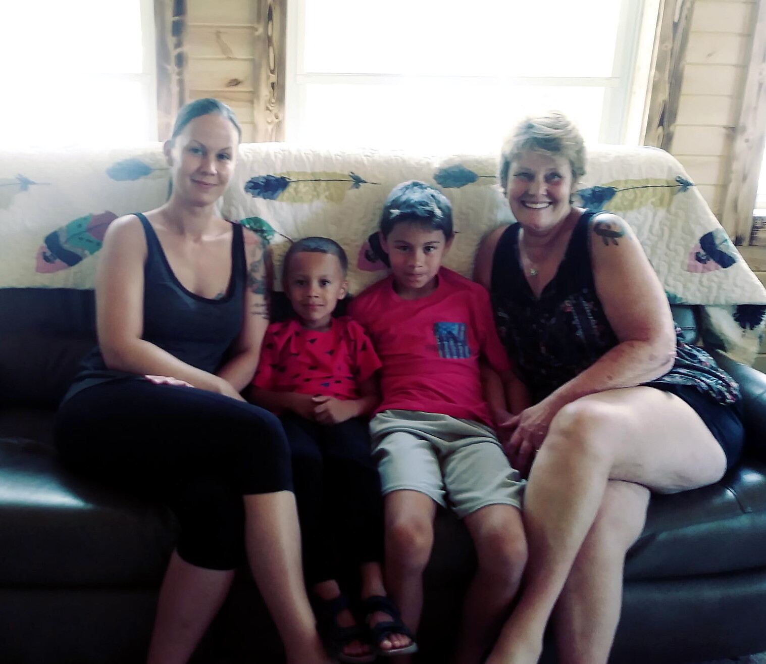 Four people—a woman on each end and two children in the middle—sitting on a sofa in a bright room, smiling at the camera.