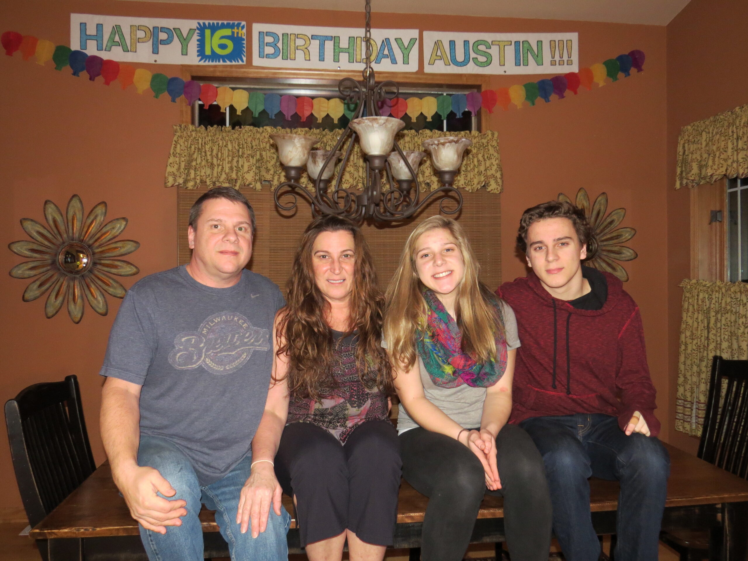 Five people are seated together on a bench in a room decorated for a birthday party, with a colorful 'Happy 16th Birthday Austin!!!' banner above them.