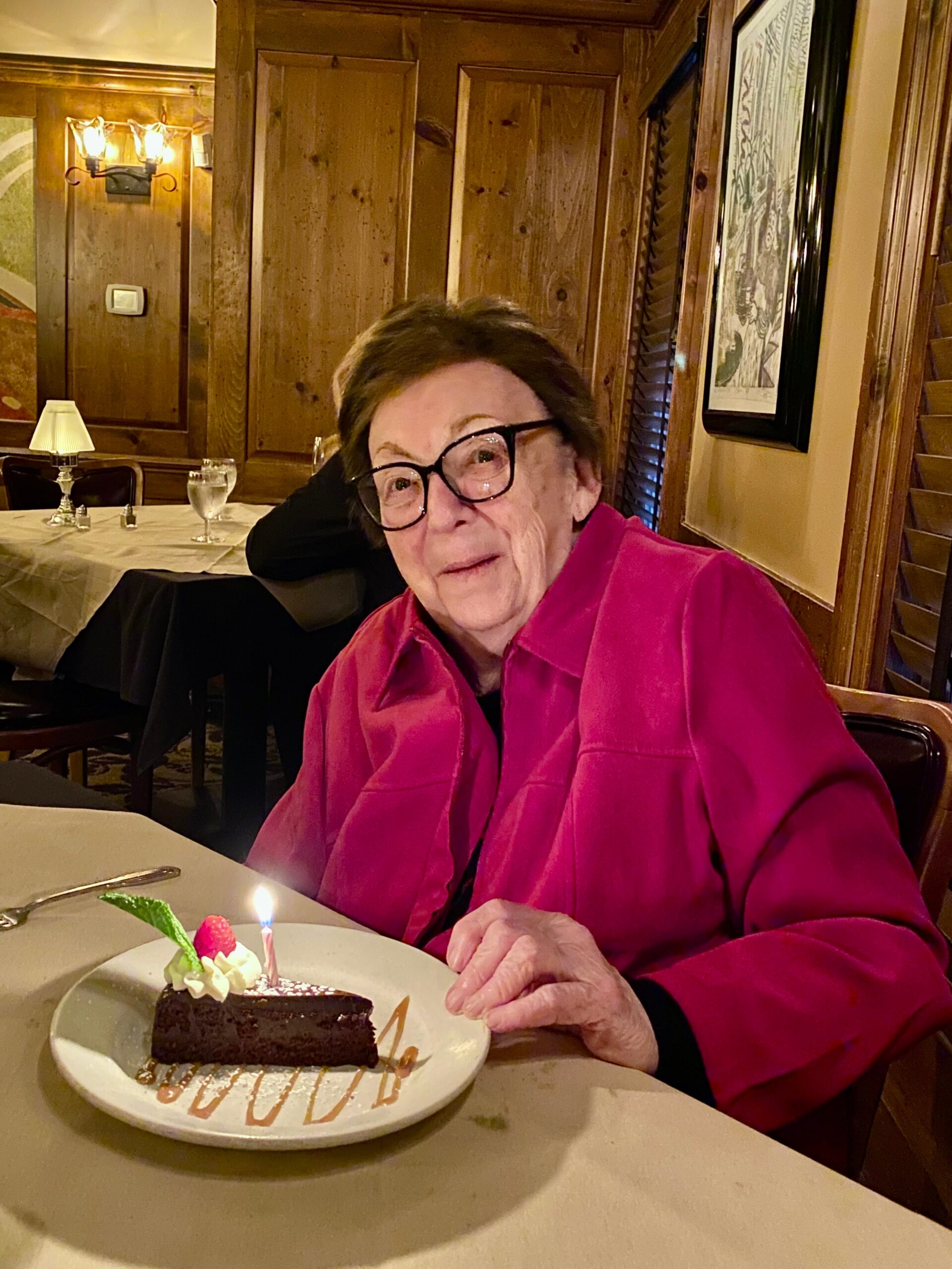 Elderly woman in a pink shirt seated at a restaurant table with a chocolate birthday cake and a lit candle in front of her.
