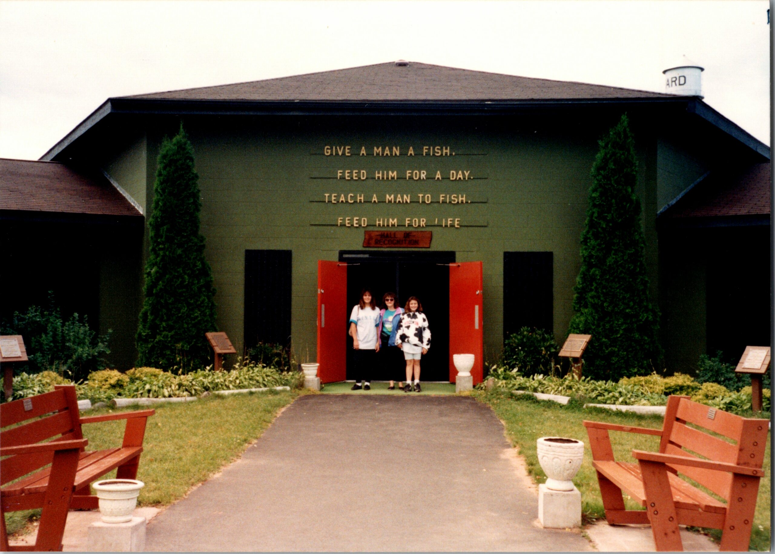 Green building facade with the quote 'GIVE A MAN A FISH... FEED HIM FOR A DAY... TEACH A MAN TO FISH... FEED HIM FOR LIFE' above a red doorway; three people stand in the entrance.