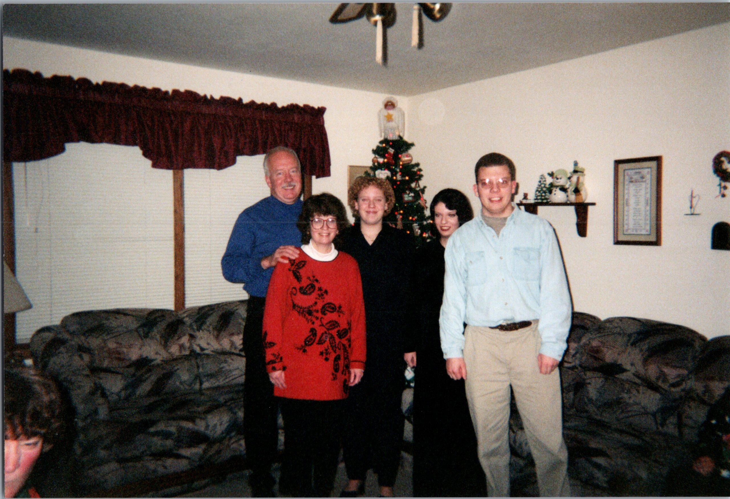 Five people pose together in a cozy living room with a Christmas tree in the background, smiling for a holiday photo (one child partially visible at the bottom).