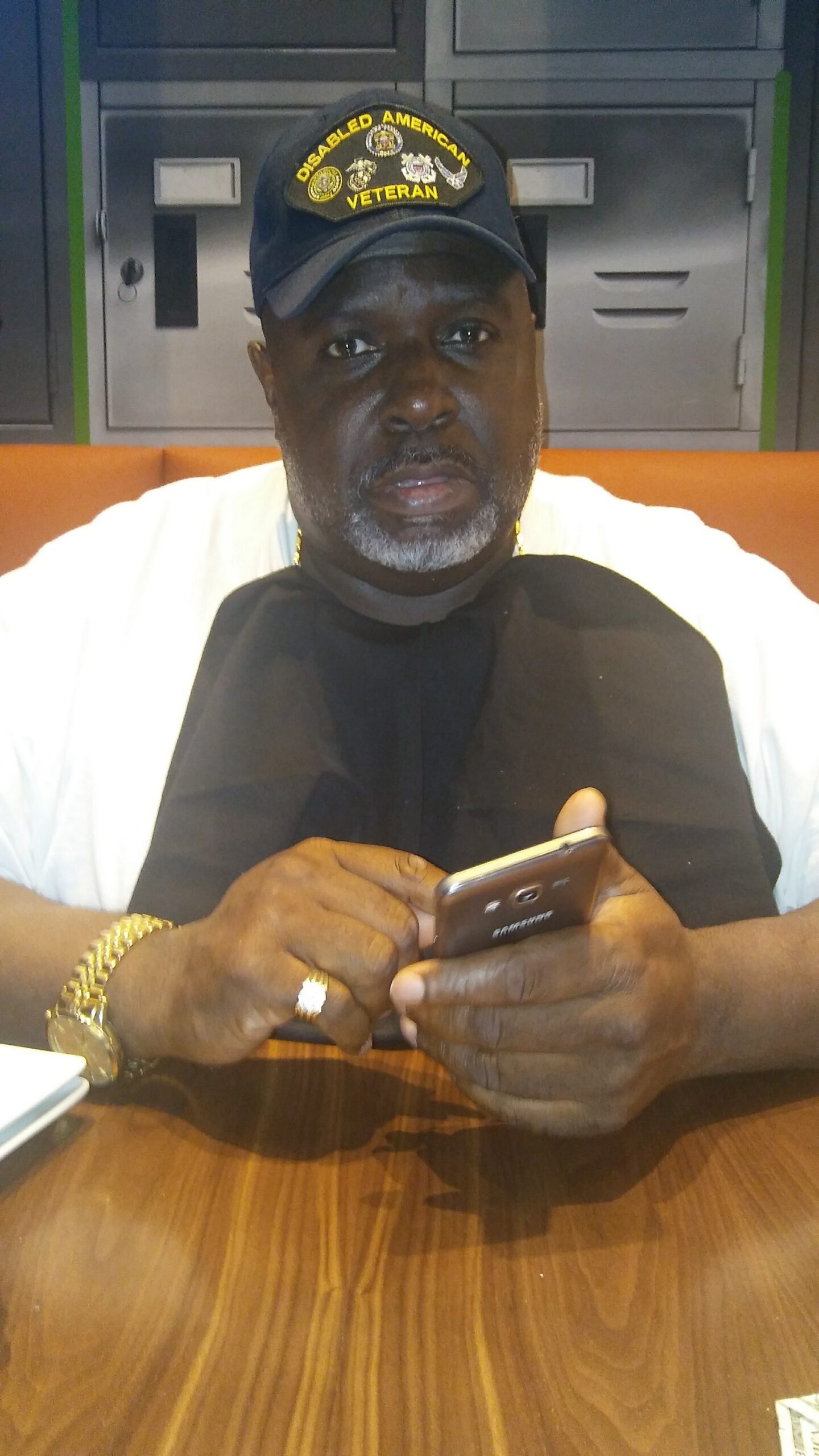 Man wearing a Disabled American Veteran cap sits at a table, looking at the camera, and holds a smartphone.