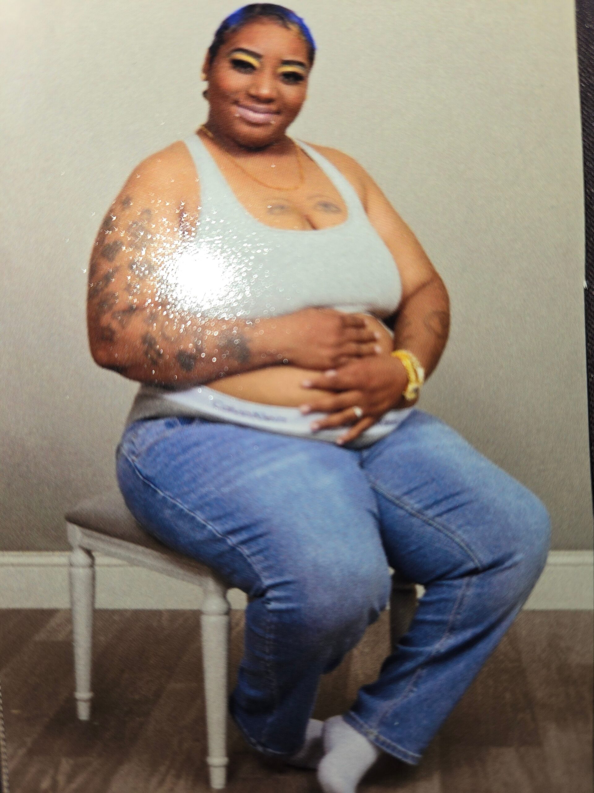 Plus-size woman seated on a white stool, wearing a gray tank top and jeans, smiling at the camera.