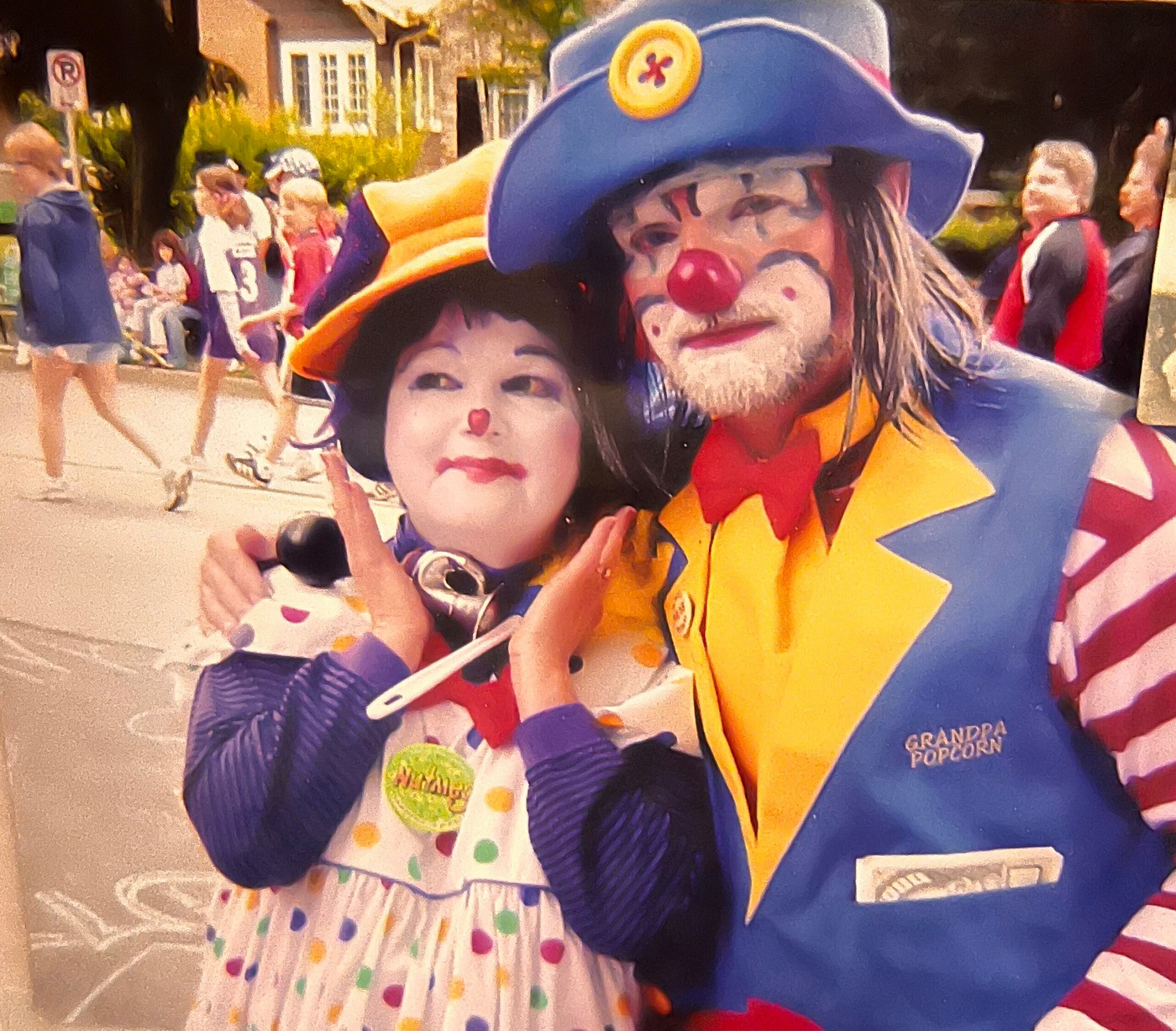 Two costumed clowns pose for a photo at an outdoor parade, with a child wearing white makeup and a red nose beside them.
