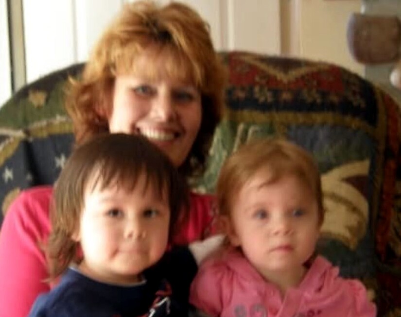 Woman with short red hair smiling on a patterned couch, holding a young boy in a navy shirt and a girl in a pink hoodie beside her at home.