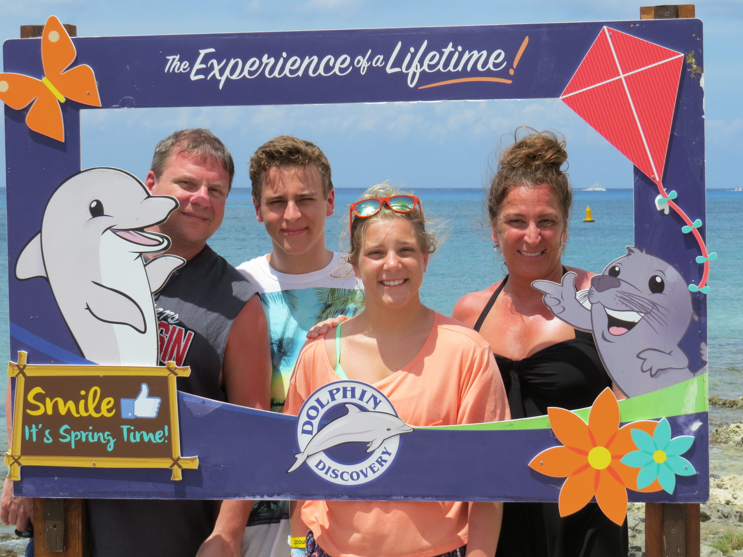 Group of four people posing behind a purple photo frame with a dolphin cutout and beach signage reading 'The Experience of a Lifetime!' by the ocean.