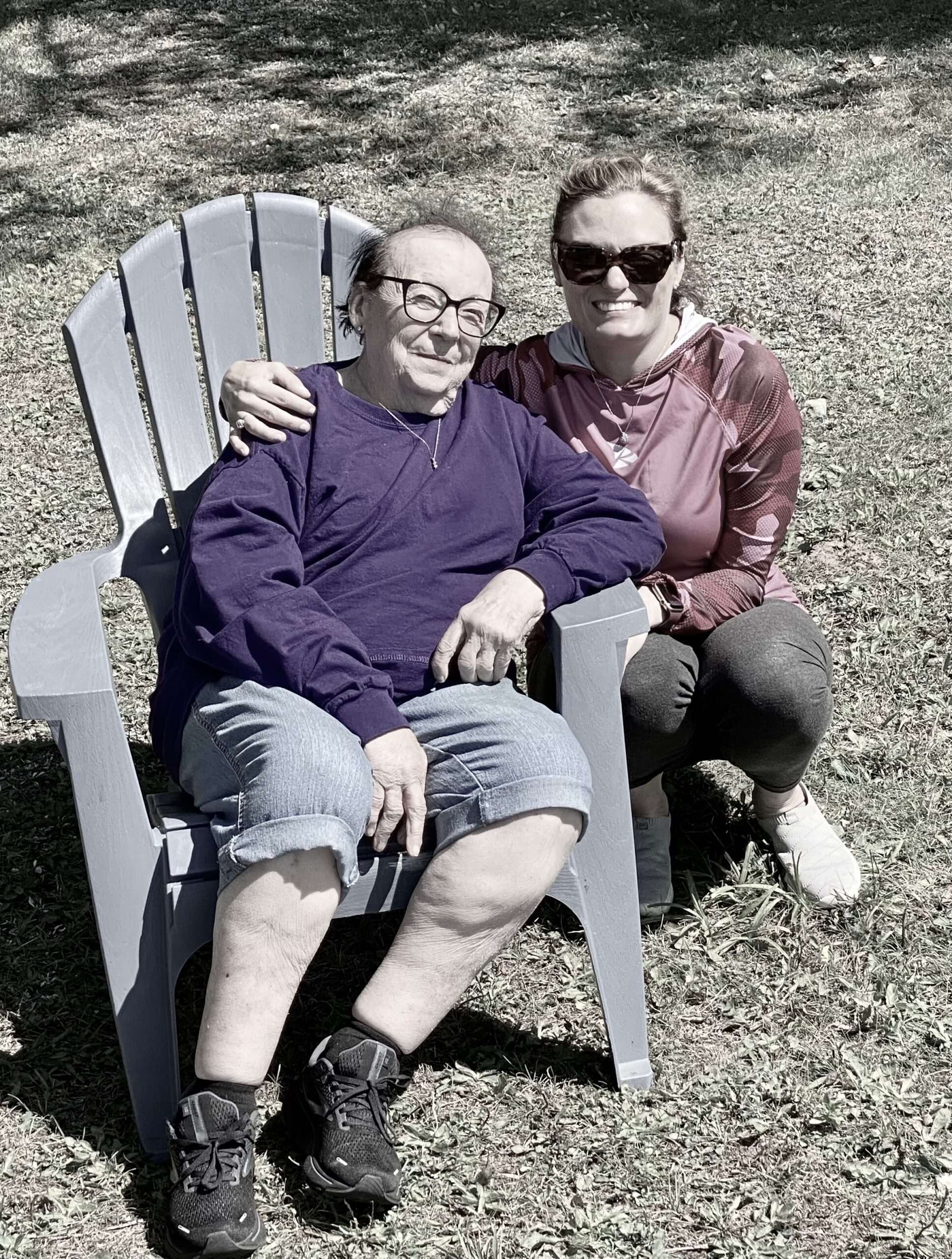 Two women sit together in a gray Adirondack chair on a sunny grassy yard, smiling at the camera with one arm around the other.