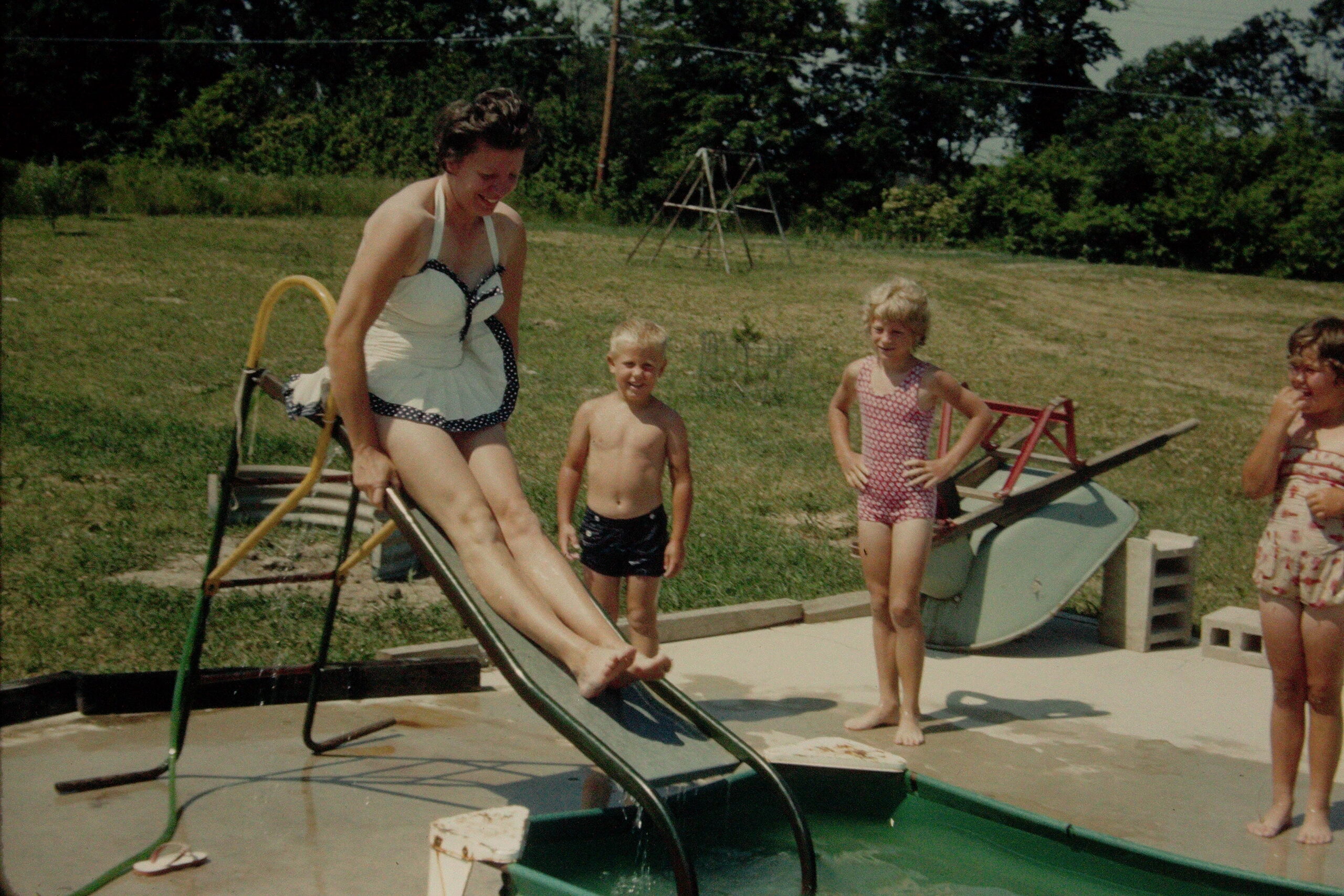 Woman in a white one-piece swimsuit slides down a metal backyard water slide as three children in swimsuits watch in a sunny grassy yard beyond a concrete patio.