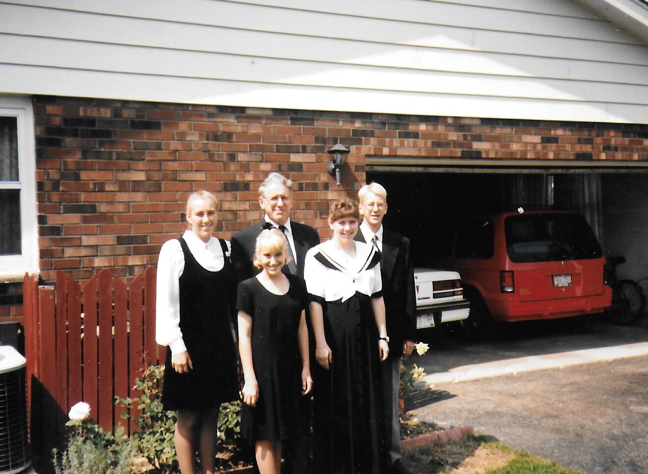 Family of five in formal black attire posing outside a brick house with a red SUV visible in the open garage.