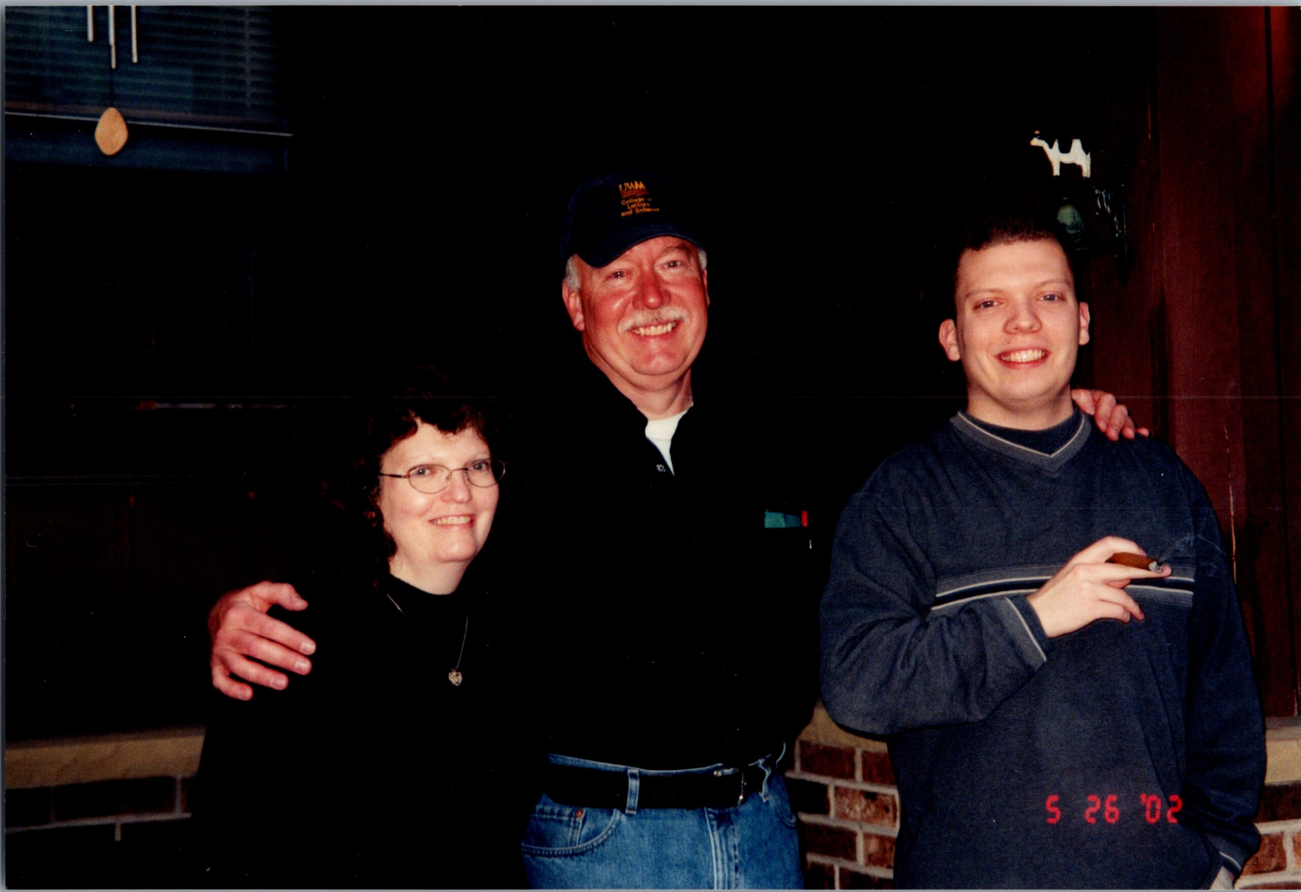 Three people posing together, an older man in a cap center, a woman with glasses on the left, and a younger man on the right, all smiling with arms around each other in a brick-walled room (date stamp 5/26/02 visible).