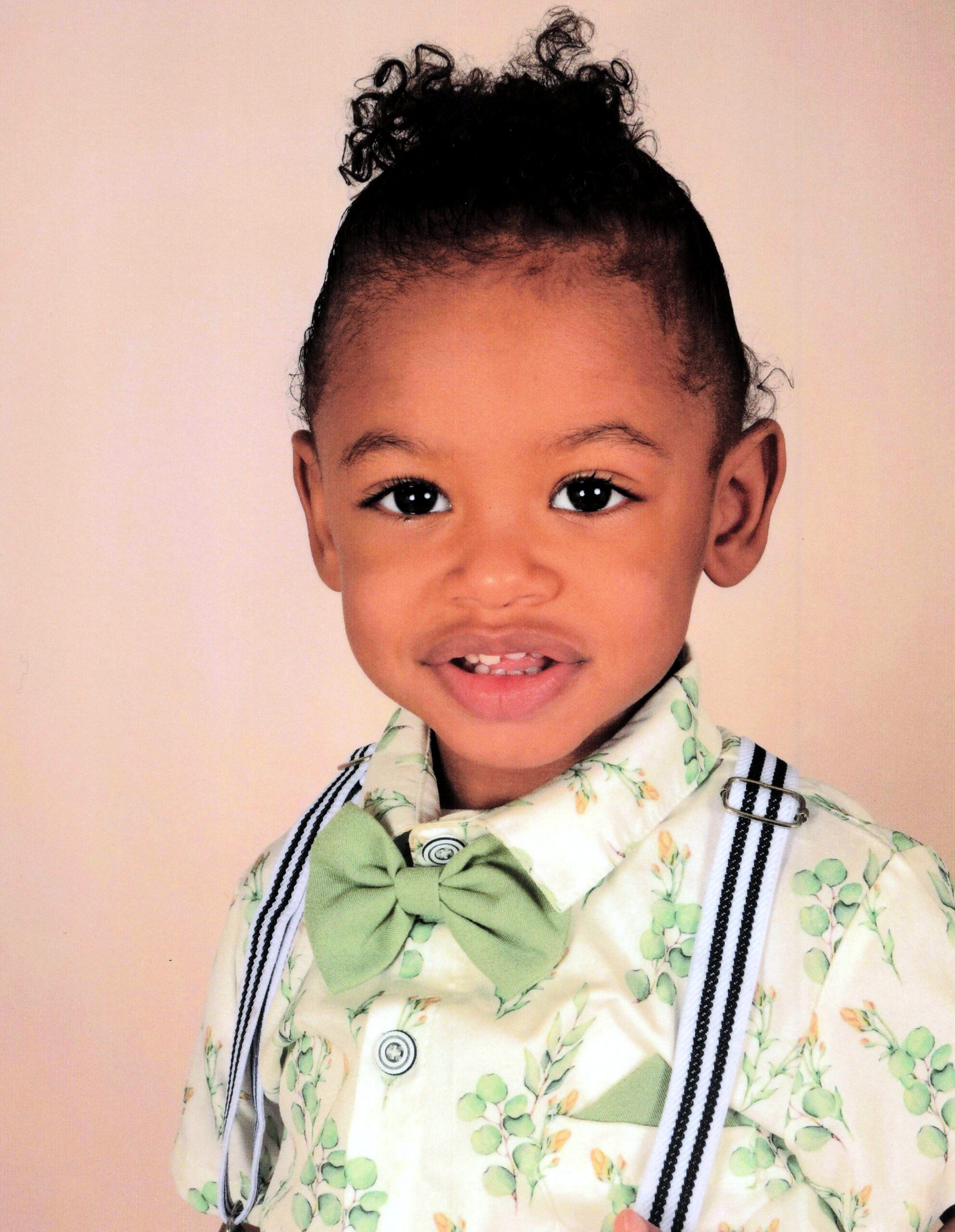 Close-up portrait of a young child with a curly hair bun, wearing a green bow tie, a floral shirt, and black-and-white suspenders against a peach background.