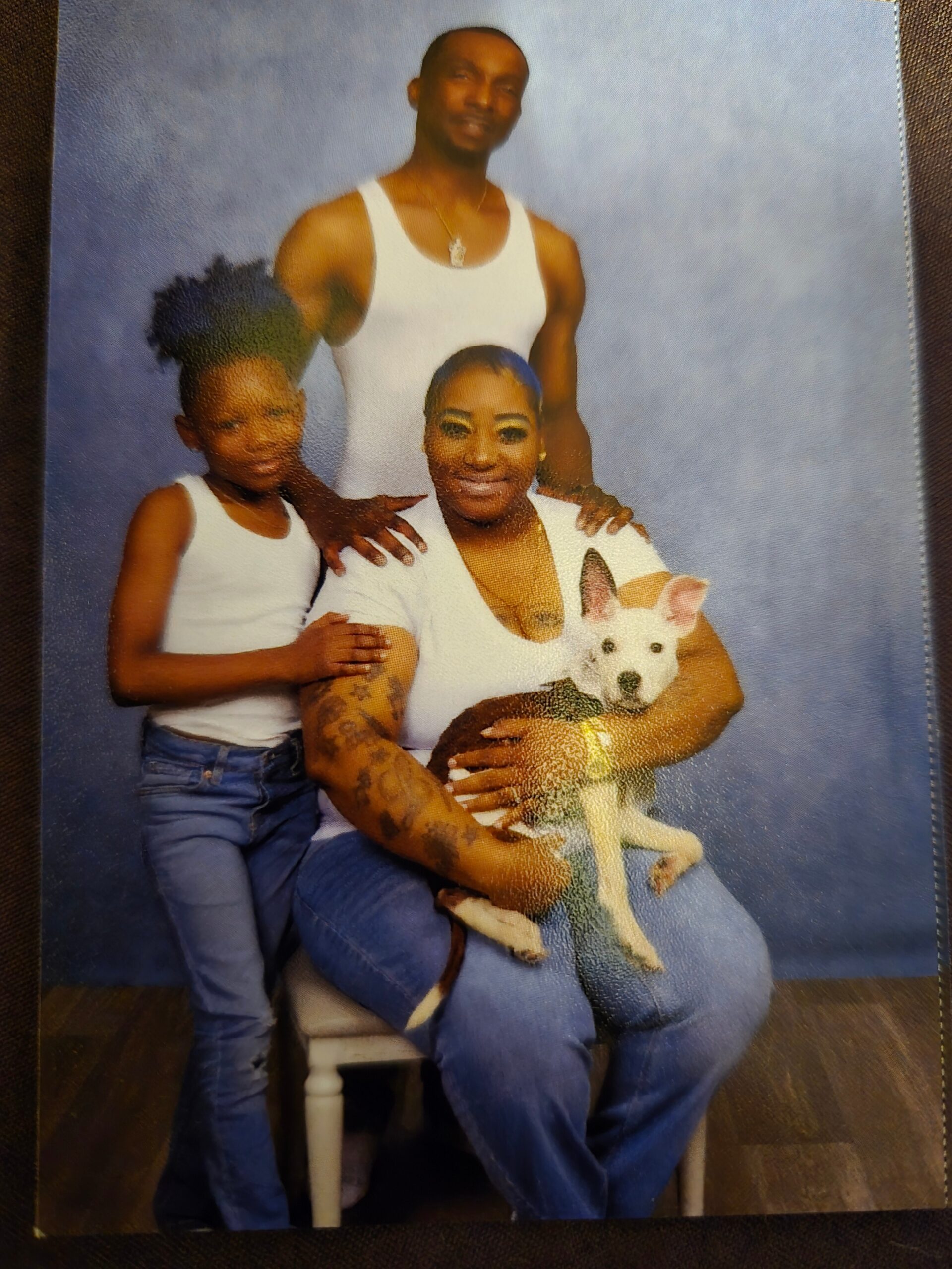 Family portrait: a man stands behind a seated woman who holds a small white dog, with a young girl beside them, all in white tops against a blue backdrop.