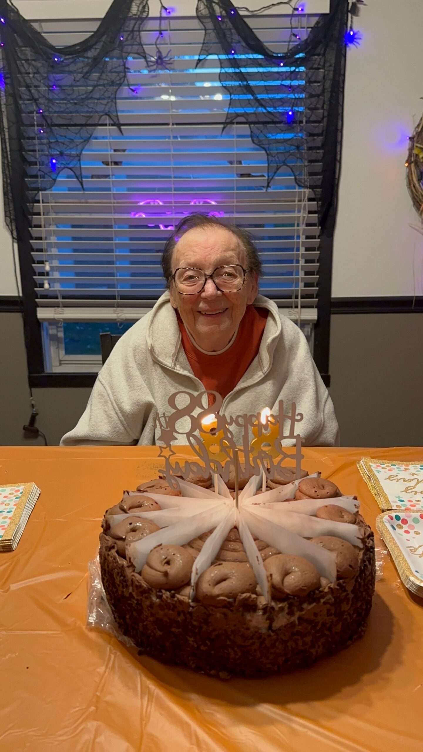 Smiling elderly person with glasses sits at a table with a chocolate birthday cake and lit candles; Halloween decorations and purple lights in the background.