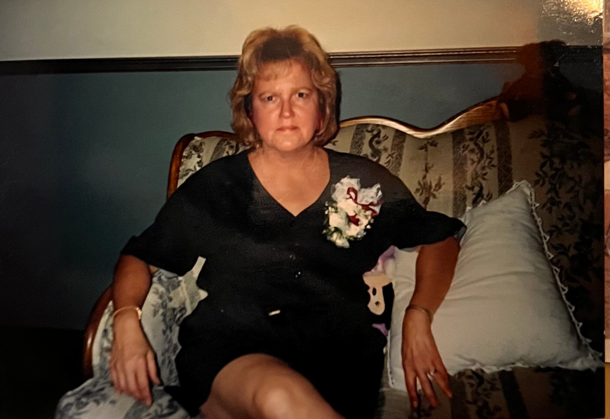 Older woman sits on a patterned sofa in a black dress with a corsage pinned to her chest.