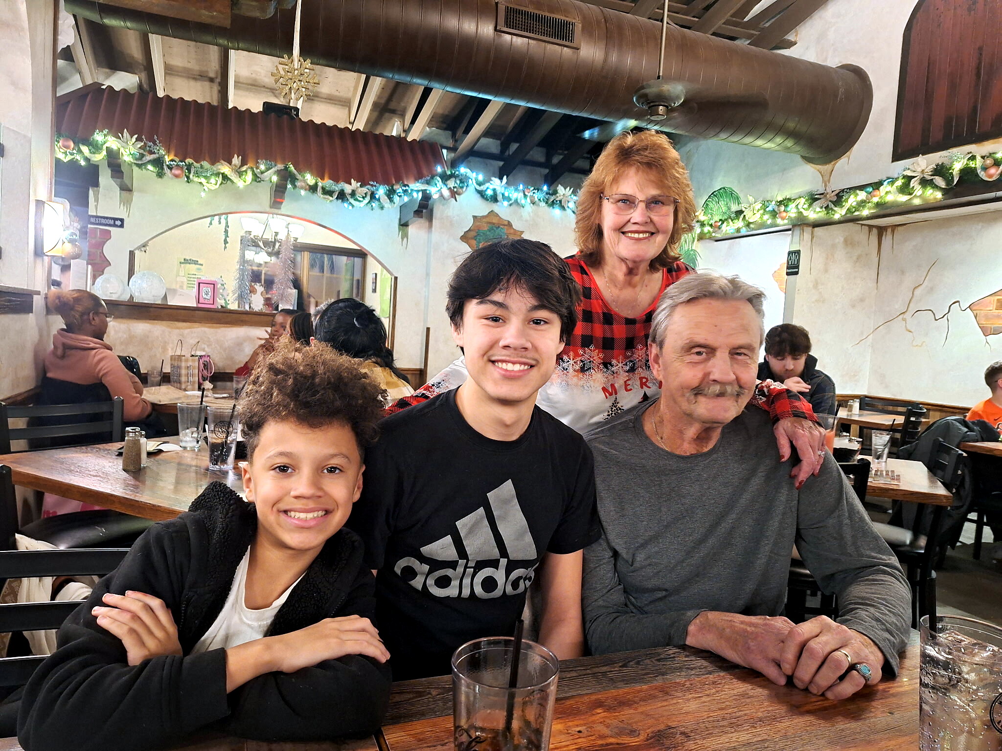 Group of people at a restaurant table smiling at the camera with festive garlands overhead and holiday decor.