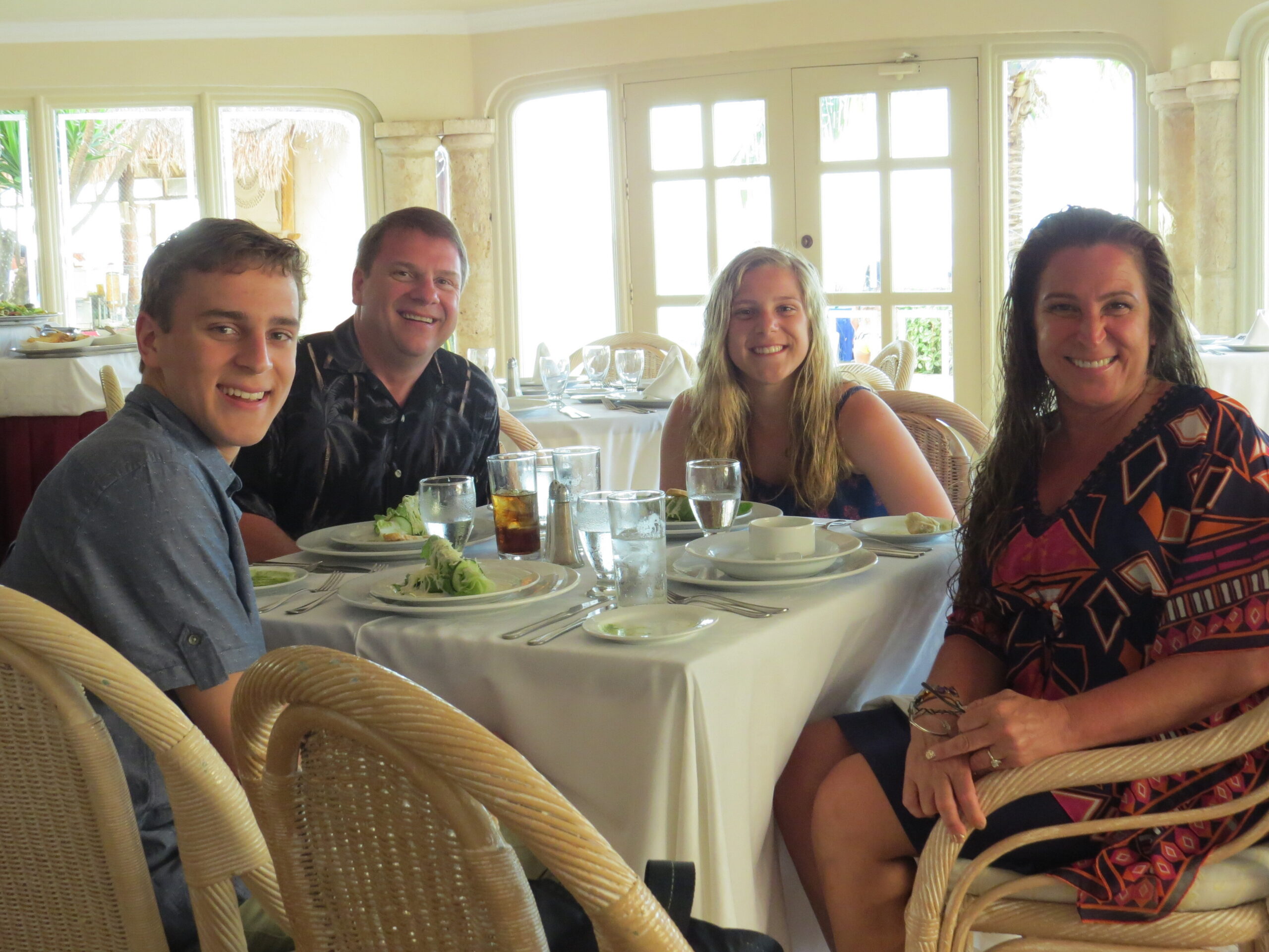 Four friends seated around a dining table, smiling at the camera in a bright restaurant setting with plates and glasses in front of them.