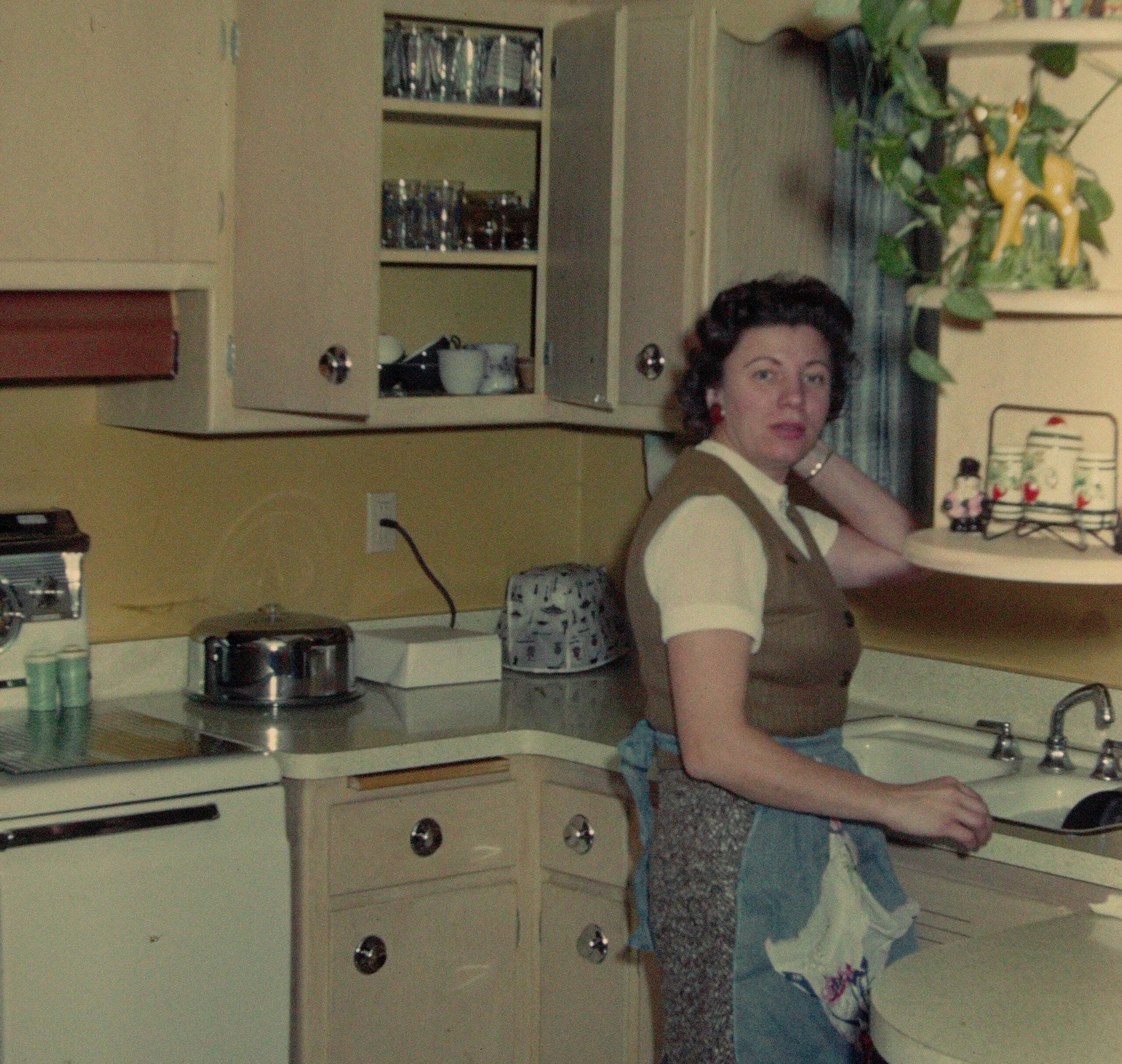 Woman stands at a vintage kitchen counter with beige cabinets and open shelves of glassware behind her.