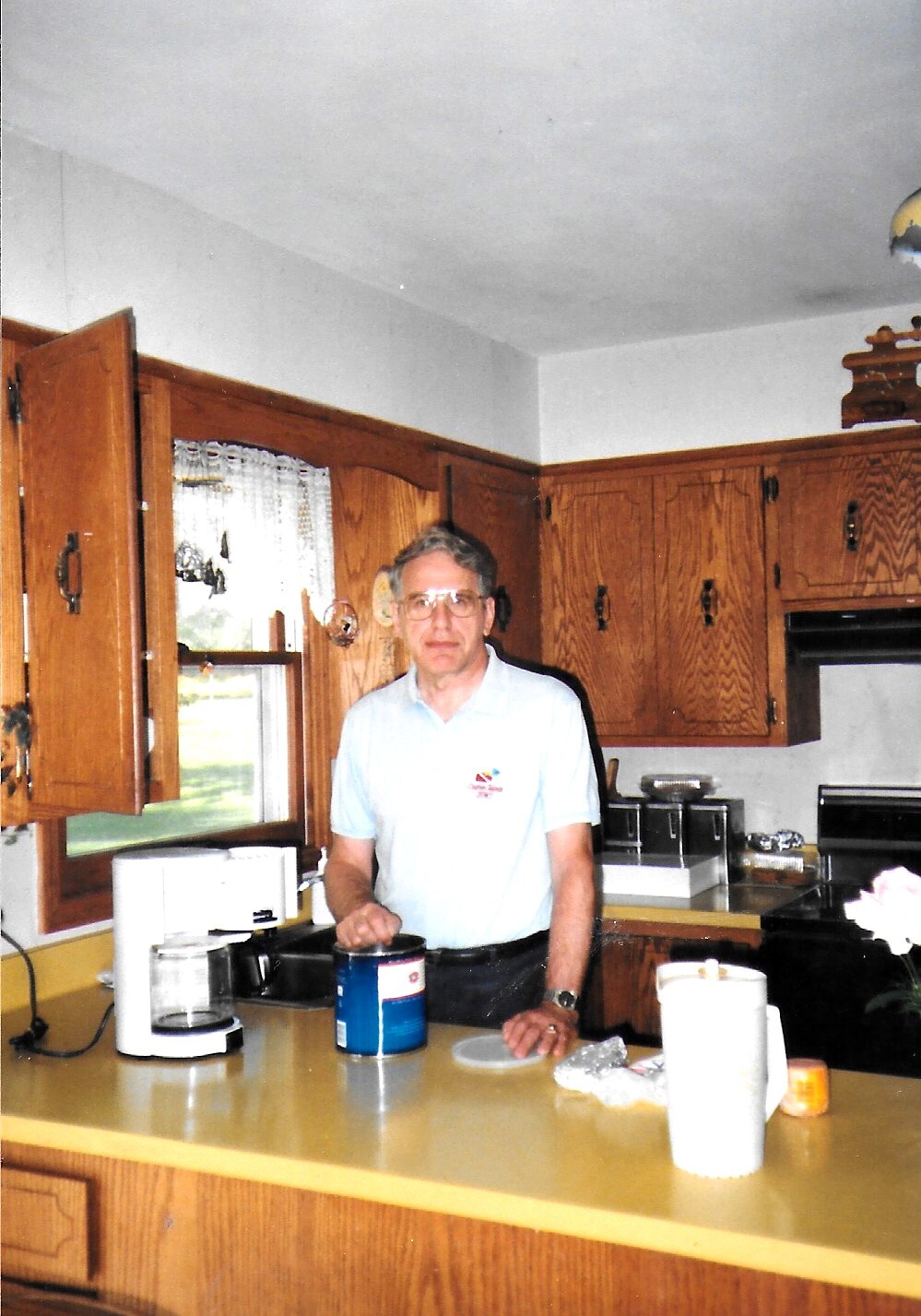 Man in a light blue polo stands at a kitchen island, handling a blue canister on the counter in front of him.