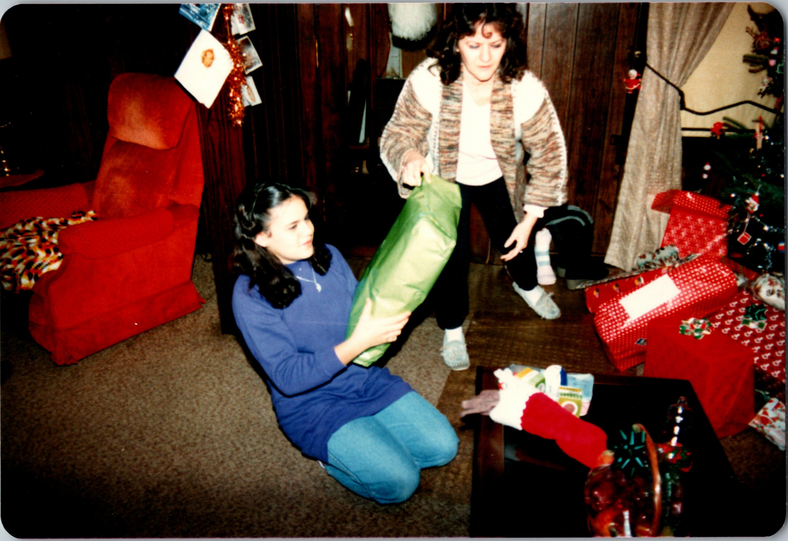 Two women exchange gifts in a cozy living room: one kneeling with a green gift bag, the other standing in a striped cardigan near a decorated Christmas tree and wrapped presents.