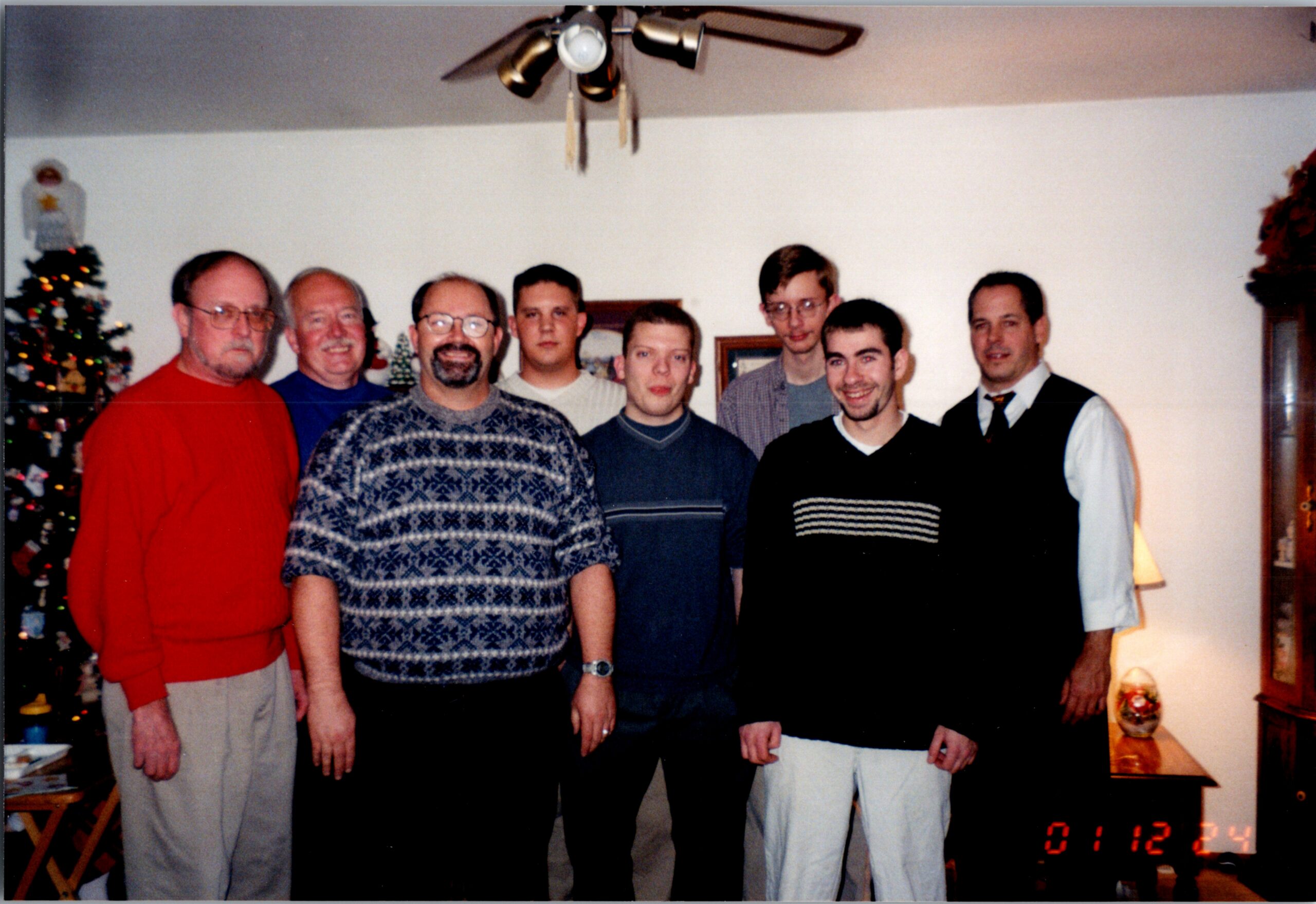 Seven men pose for a casual group photo in a living room, Christmas tree on the left and a ceiling fan overhead.
