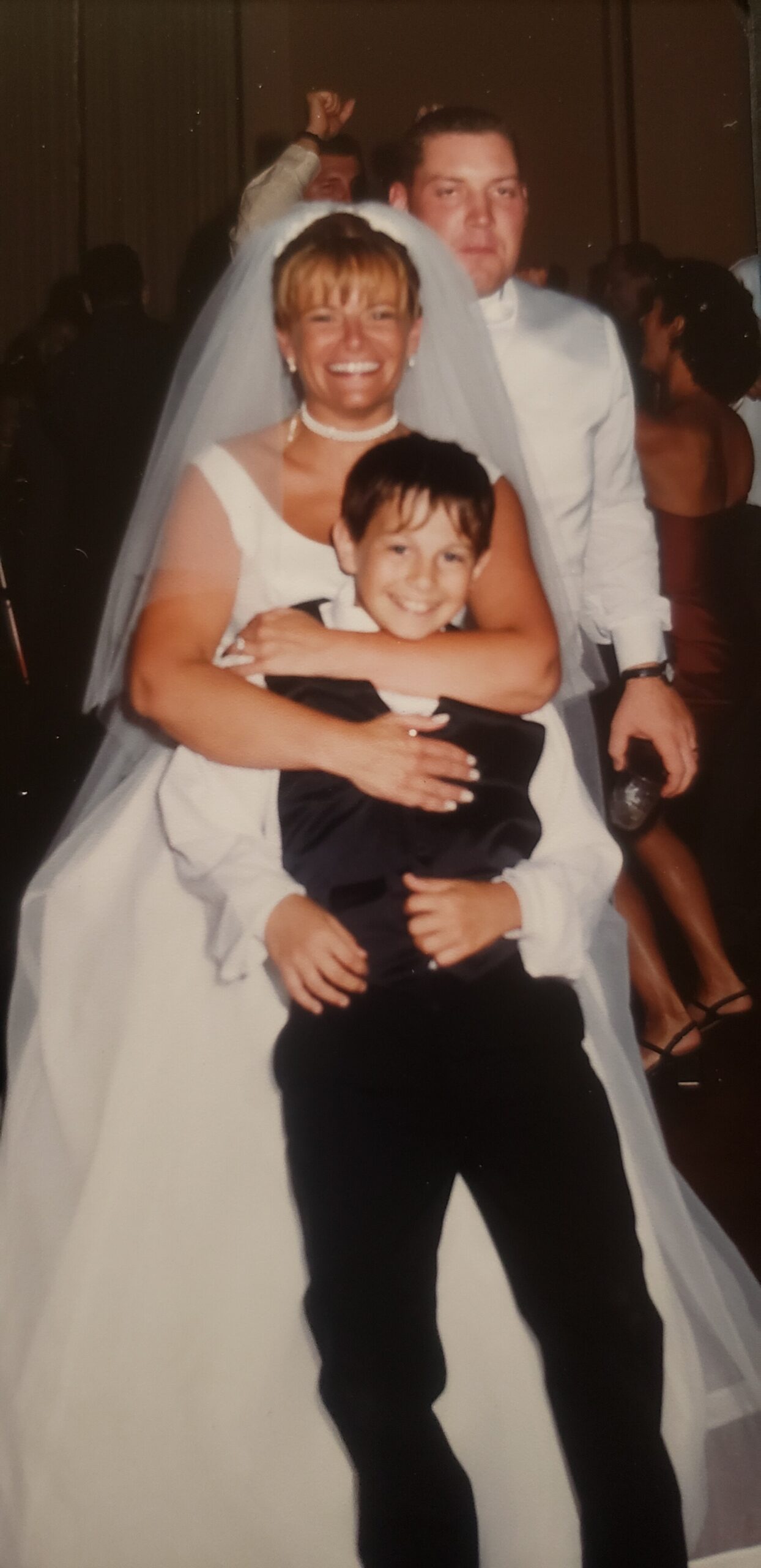 Bride in white gown and veil smiles while holding a young boy in a suit at a wedding reception.