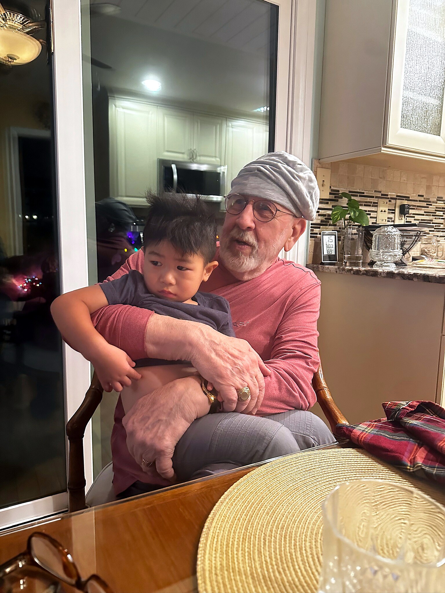 Elderly man with a gray turban and glasses holding a young child on his lap at a kitchen table.