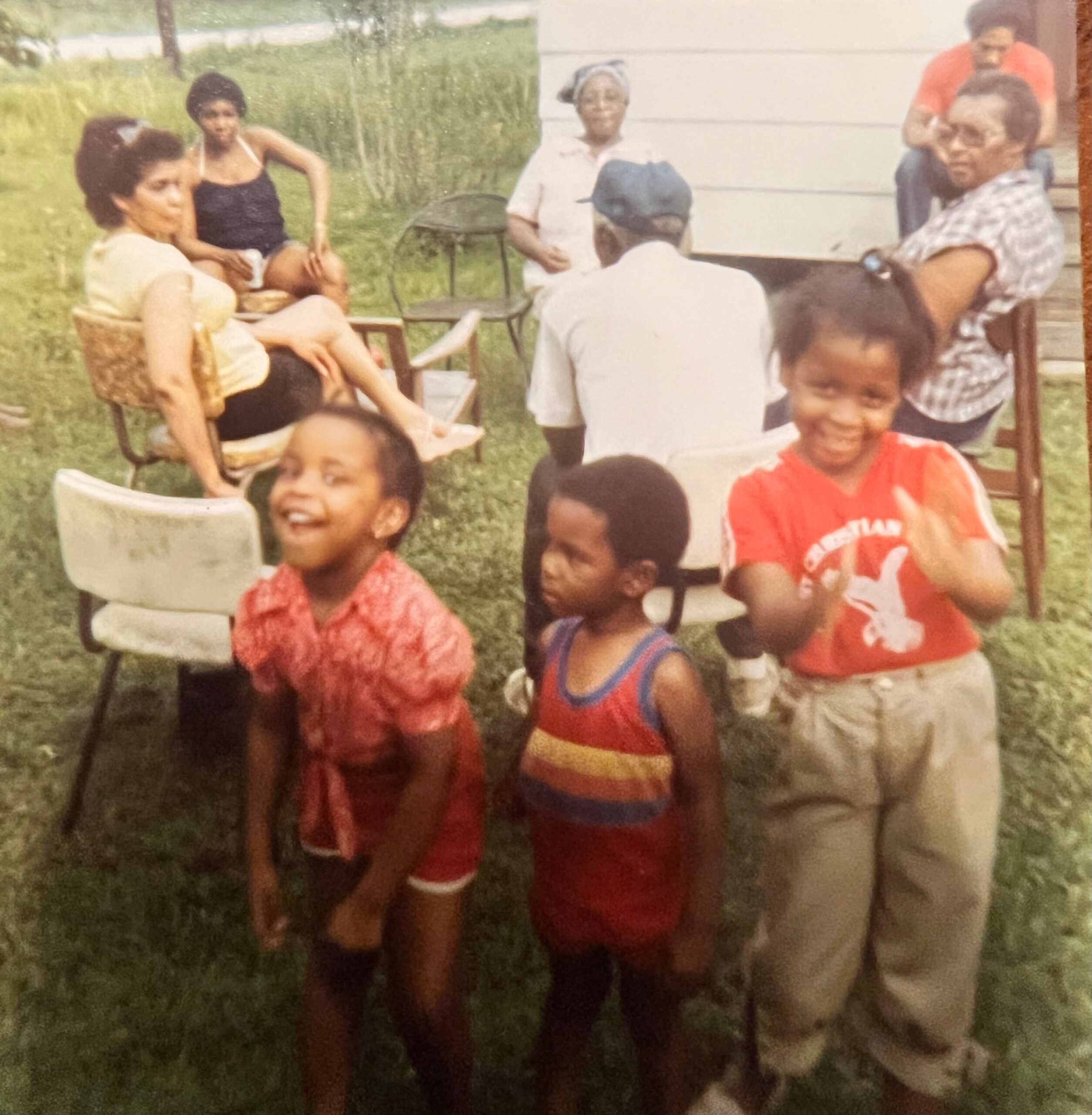 A group of family members outdoors in a grassy yard, some sitting on chairs and others standing, smiling and chatting.