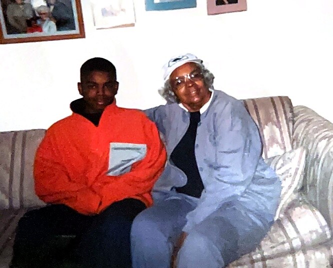 Two people sit closely on a striped couch: a young man in an orange jacket and an older woman in a light blue outfit with a white headscarf.